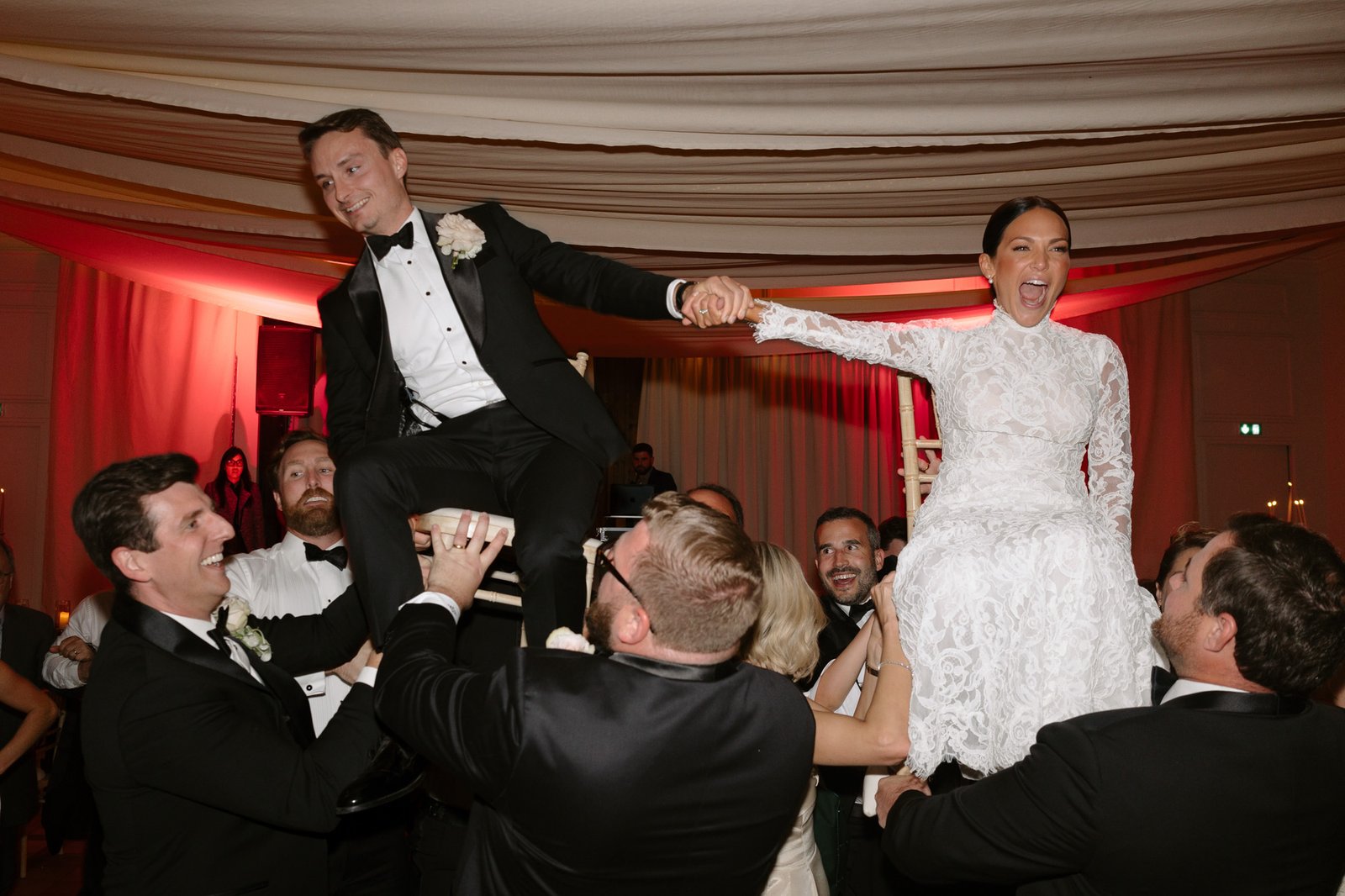 A bride and groom are lifted on chairs by guests during a wedding celebration, holding hands and smiling under draped fabric. Chateau de la Valouze wedding.