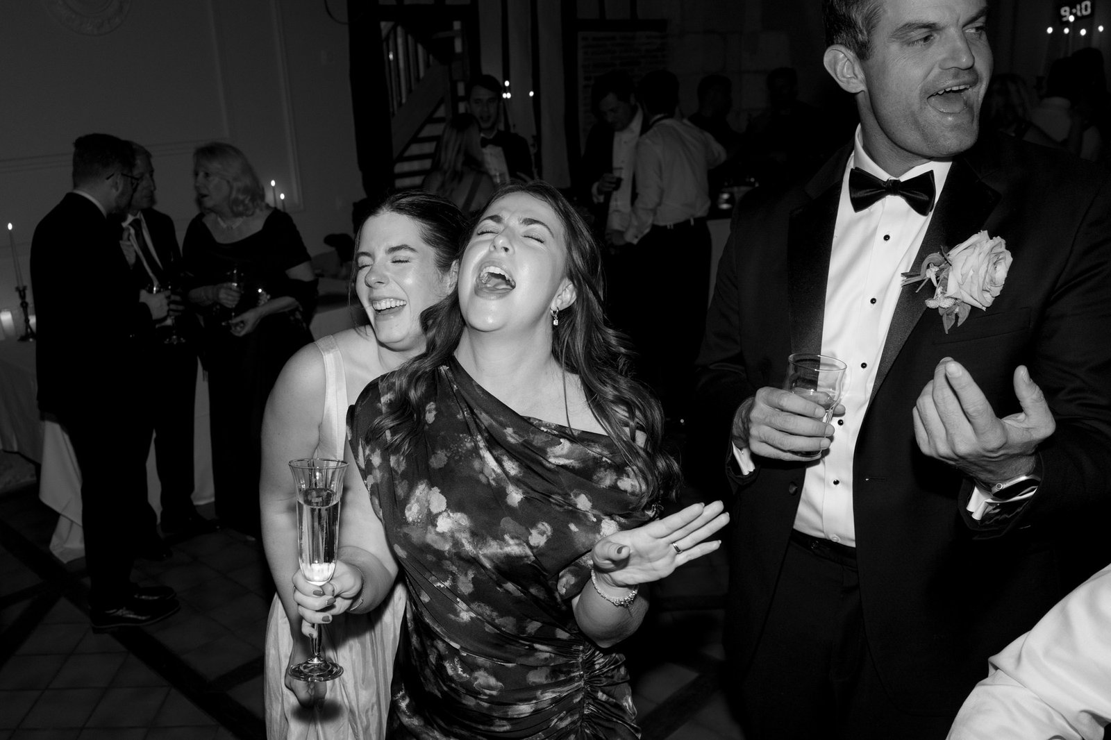 Three people, dressed formally, sing and laugh at a lively indoor event. Others in the background mingle and hold drinks. The scene appears to be at a celebration or party.