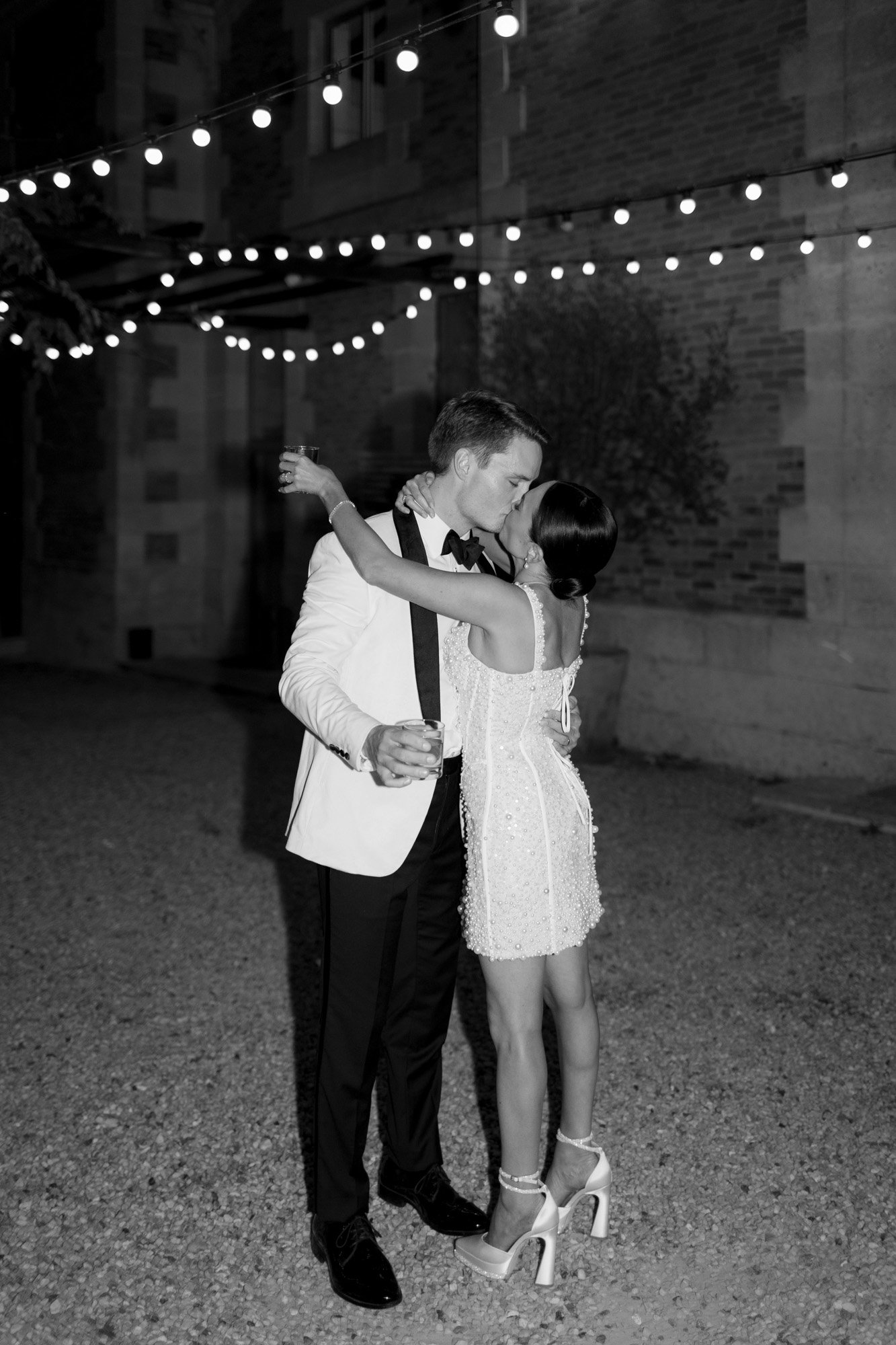 A couple dressed in formal attire embraces under string lights at night; the man holds a drink and the woman wears high heels and a short dress. Chateau de la Valouze wedding.