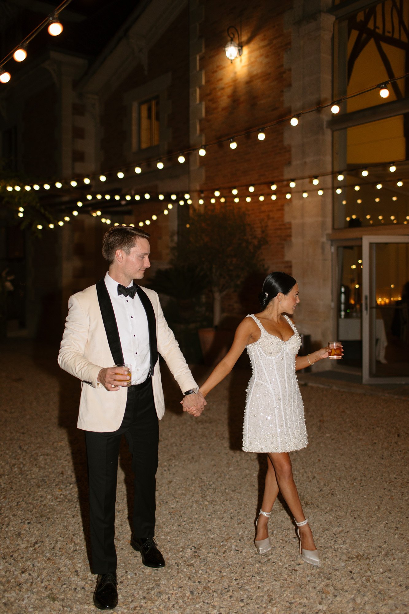 A man in a white tuxedo jacket and a woman in a short white dress hold hands and drinks while walking outside at night under string lights. Chateau de la Valouze wedding.