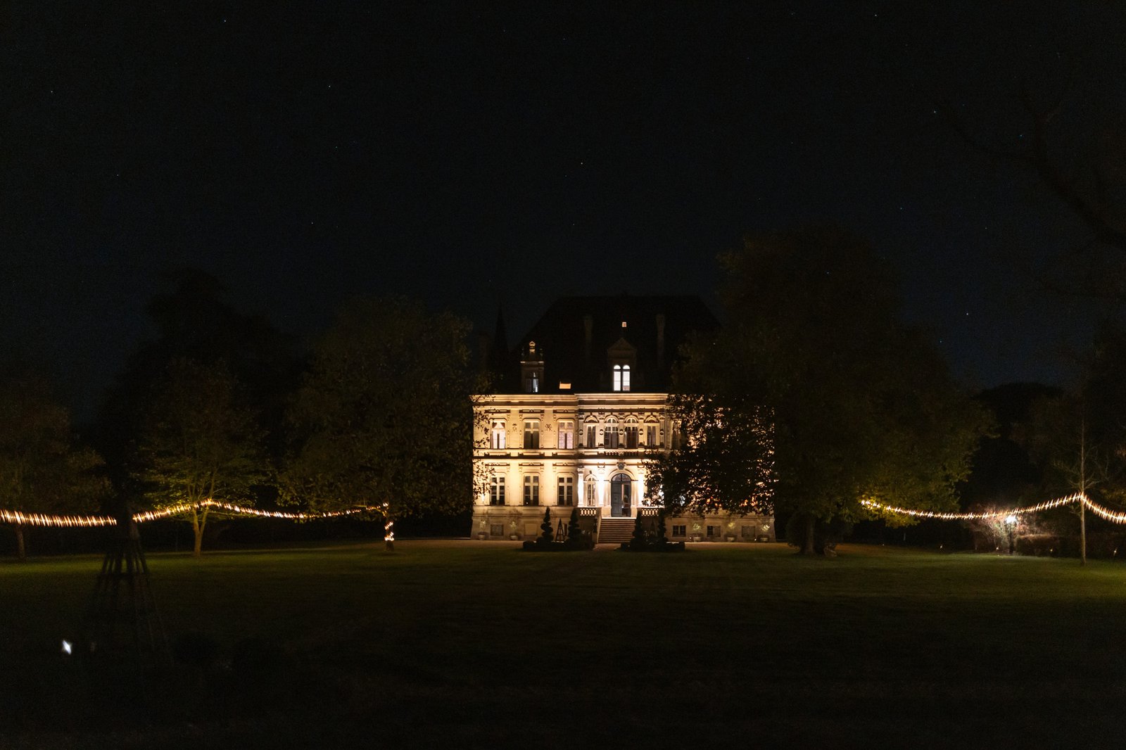 A large, historic building is illuminated at night, surrounded by trees and string lights on a spacious lawn. Chateau de la Valouze wedding.