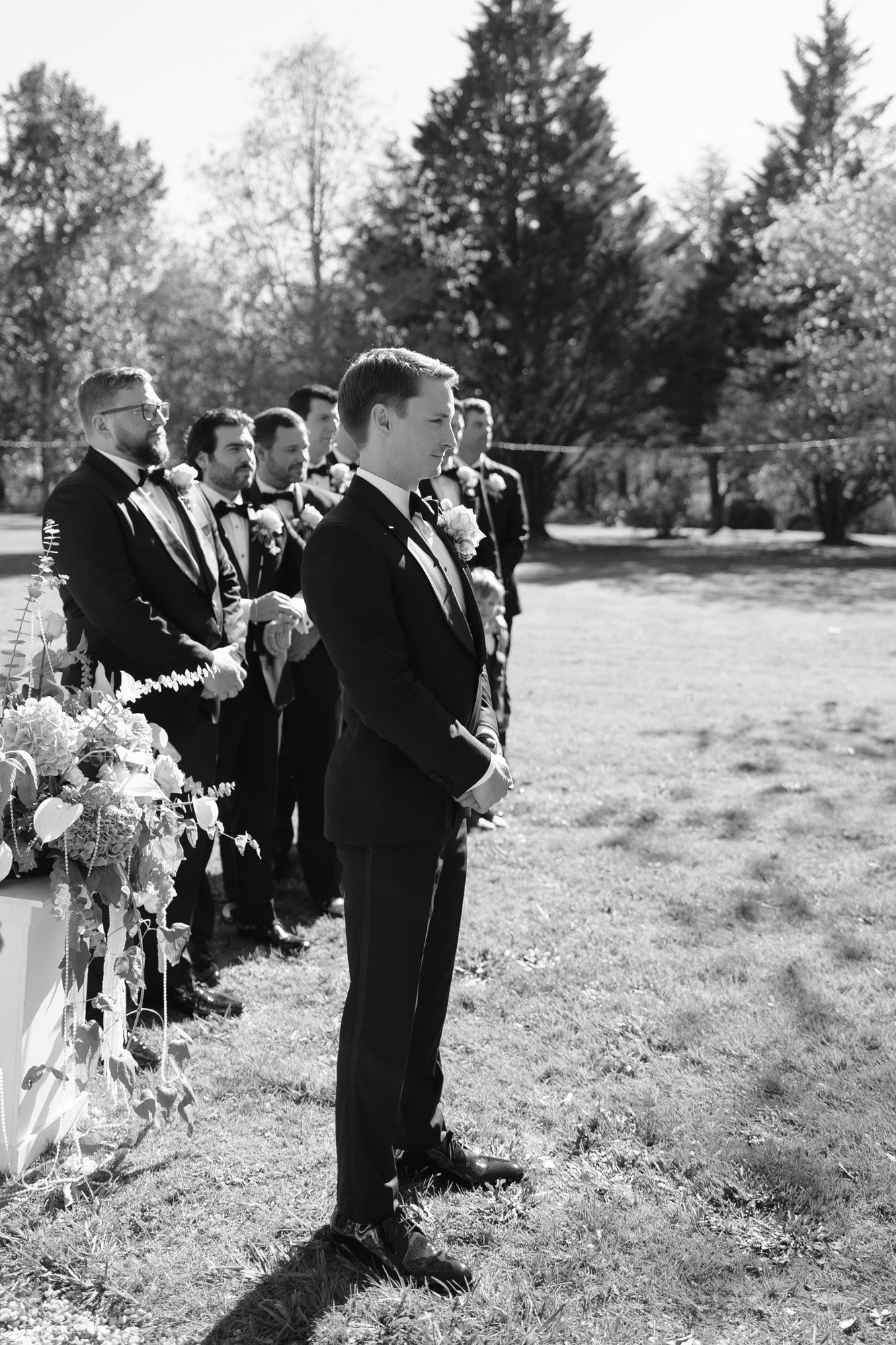 A group of men in suits, including one standing at the front, stand outdoors during a formal event with trees and flowers in the background.