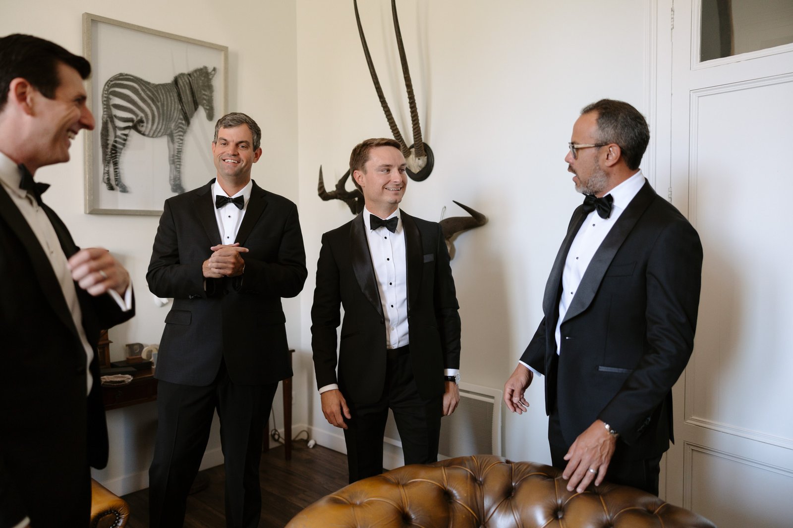 Four men in black tuxedos stand and talk in a room decorated with animal art and horns, appearing to prepare for a formal event. Chateau de la Valouze wedding.