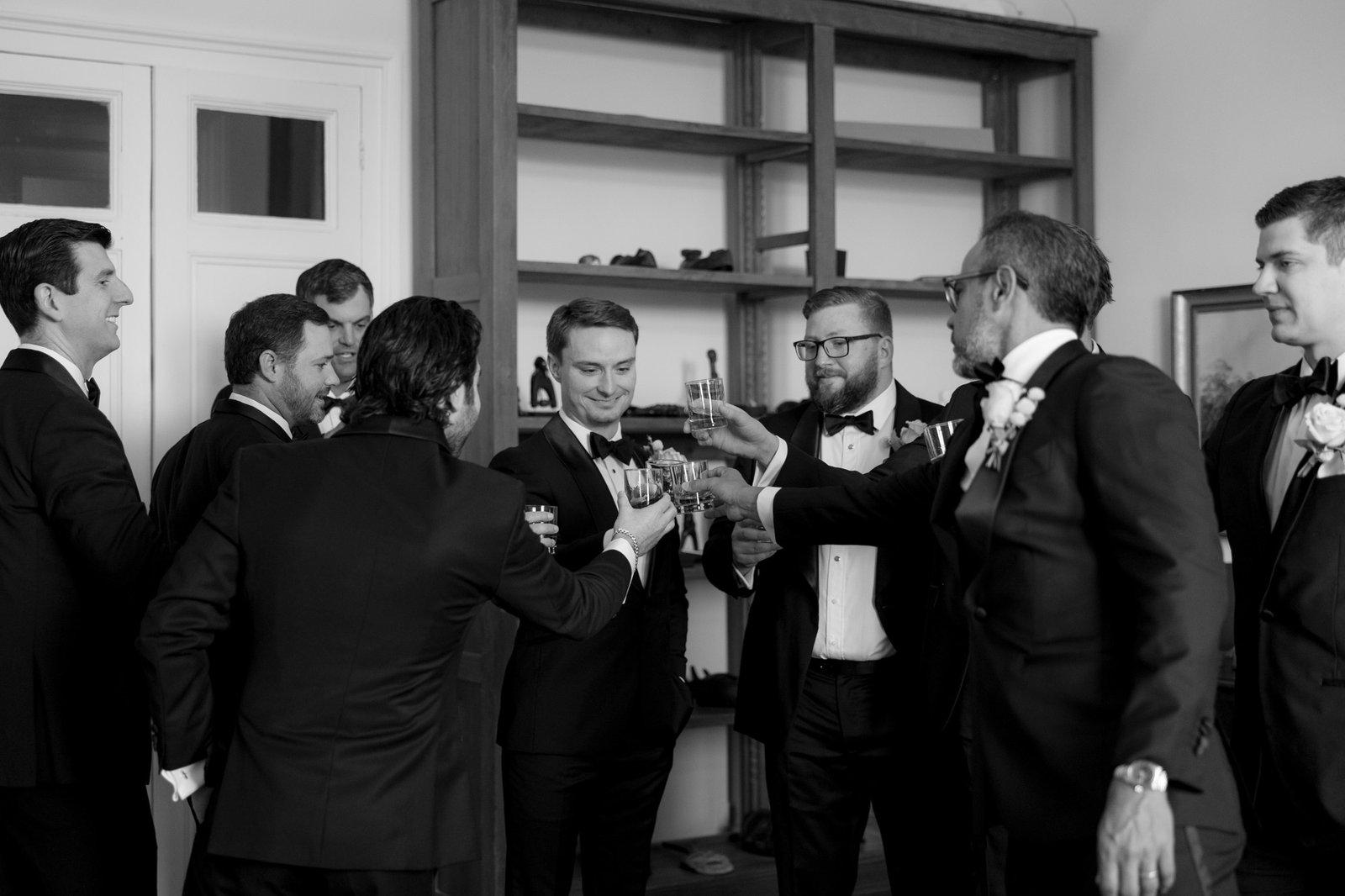 Seven men in suits and tuxedos stand together indoors, raising glasses in a toast. The scene appears formal, with smiles and shelves in the background. The image is in black and white. Chateau de la Valouze wedding.