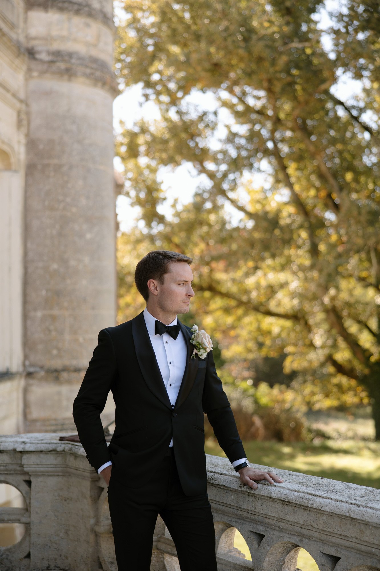 A man in a black tuxedo with a boutonniere stands by a stone railing outdoors, with trees and a historic building in the background.