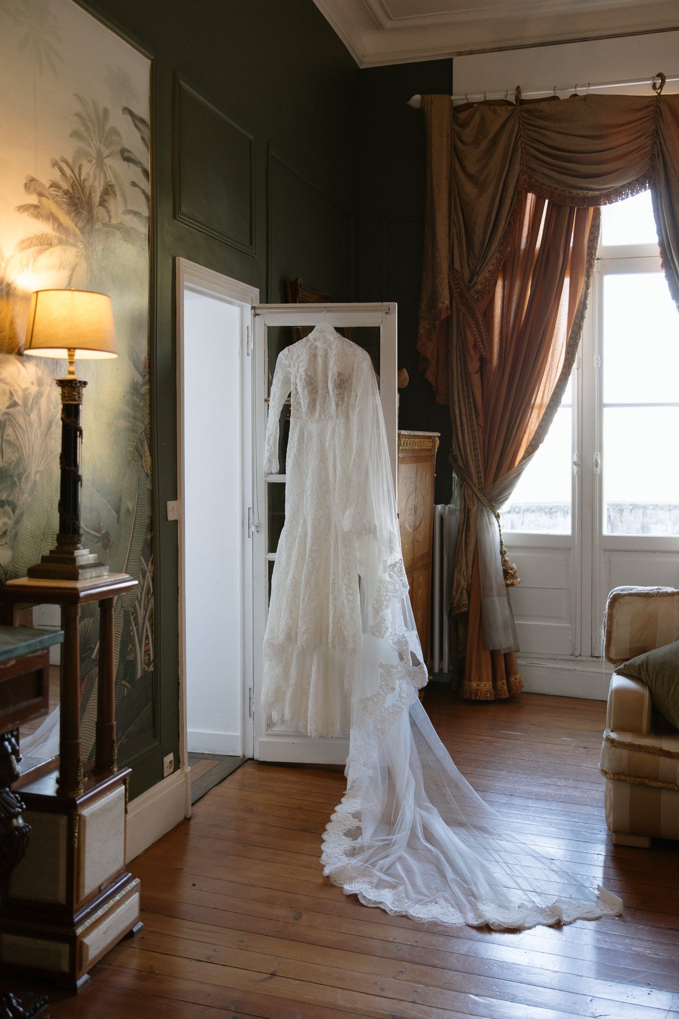 A long-sleeved white lace wedding dress with a train hangs on a glass door in an elegant, vintage-style room with wooden floors and ornate curtains. Chateau de la Valouze wedding.
