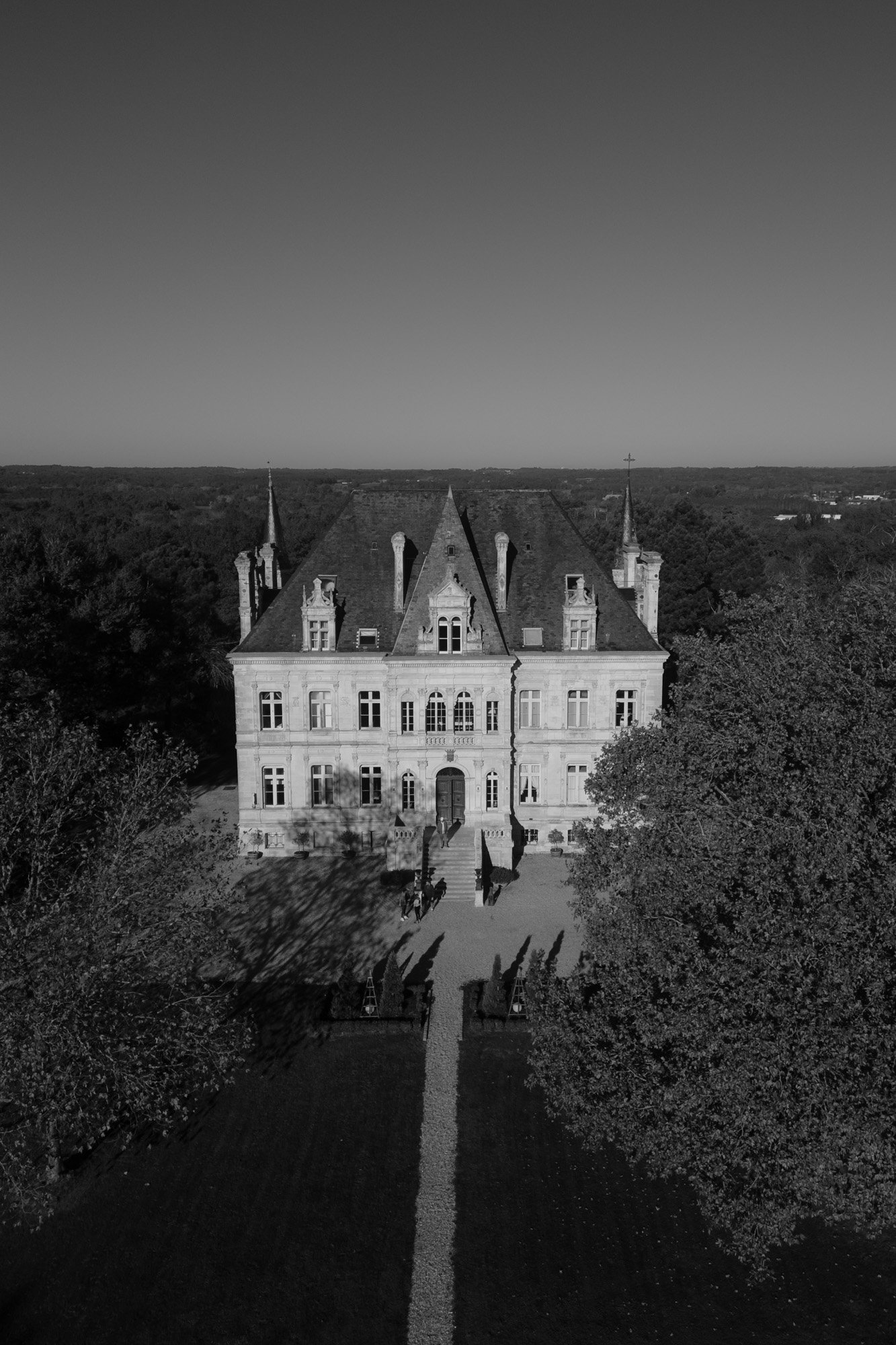 Aerial view of a large, historic stone mansion with steep roofs, surrounded by trees and a long pathway leading to the entrance, photographed in black and white. Chateau de la Valouze wedding.