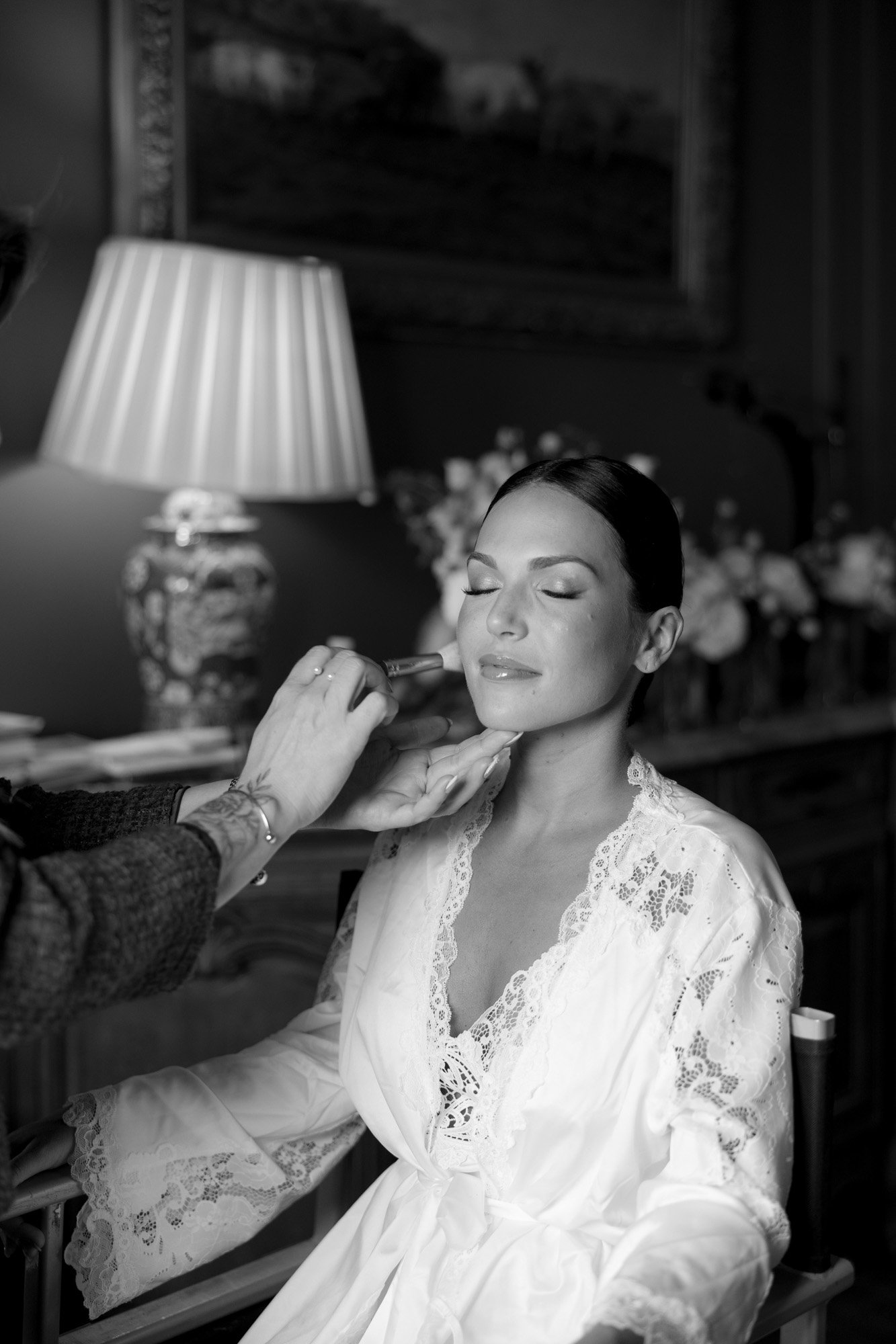 A woman in a lace robe sits with her eyes closed while someone applies makeup to her face in a softly lit room. Chateau de la Valouze wedding.