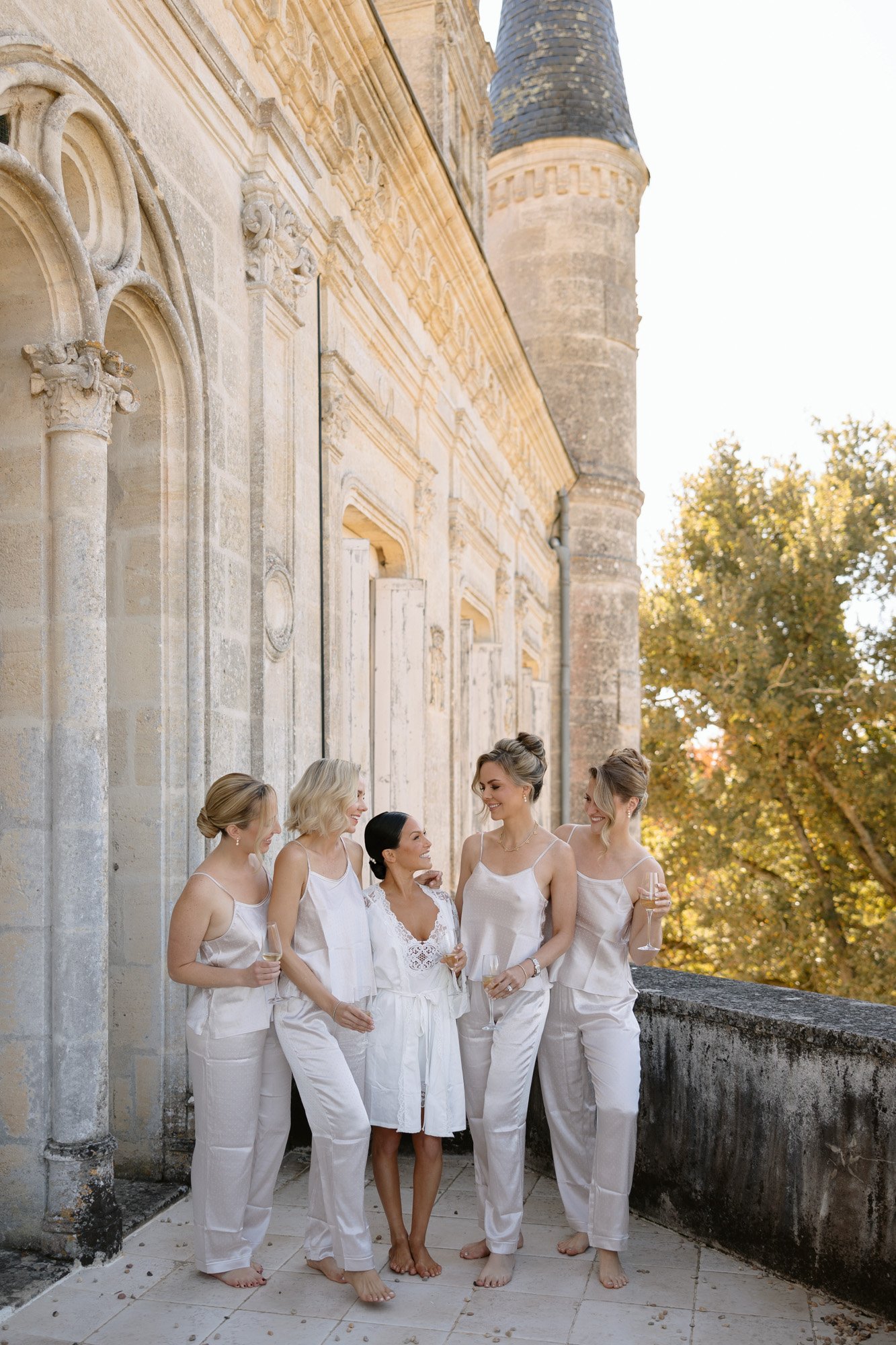 Five women in light-colored sleepwear stand and chat on a stone terrace beside a historic building with arched windows and a turret.