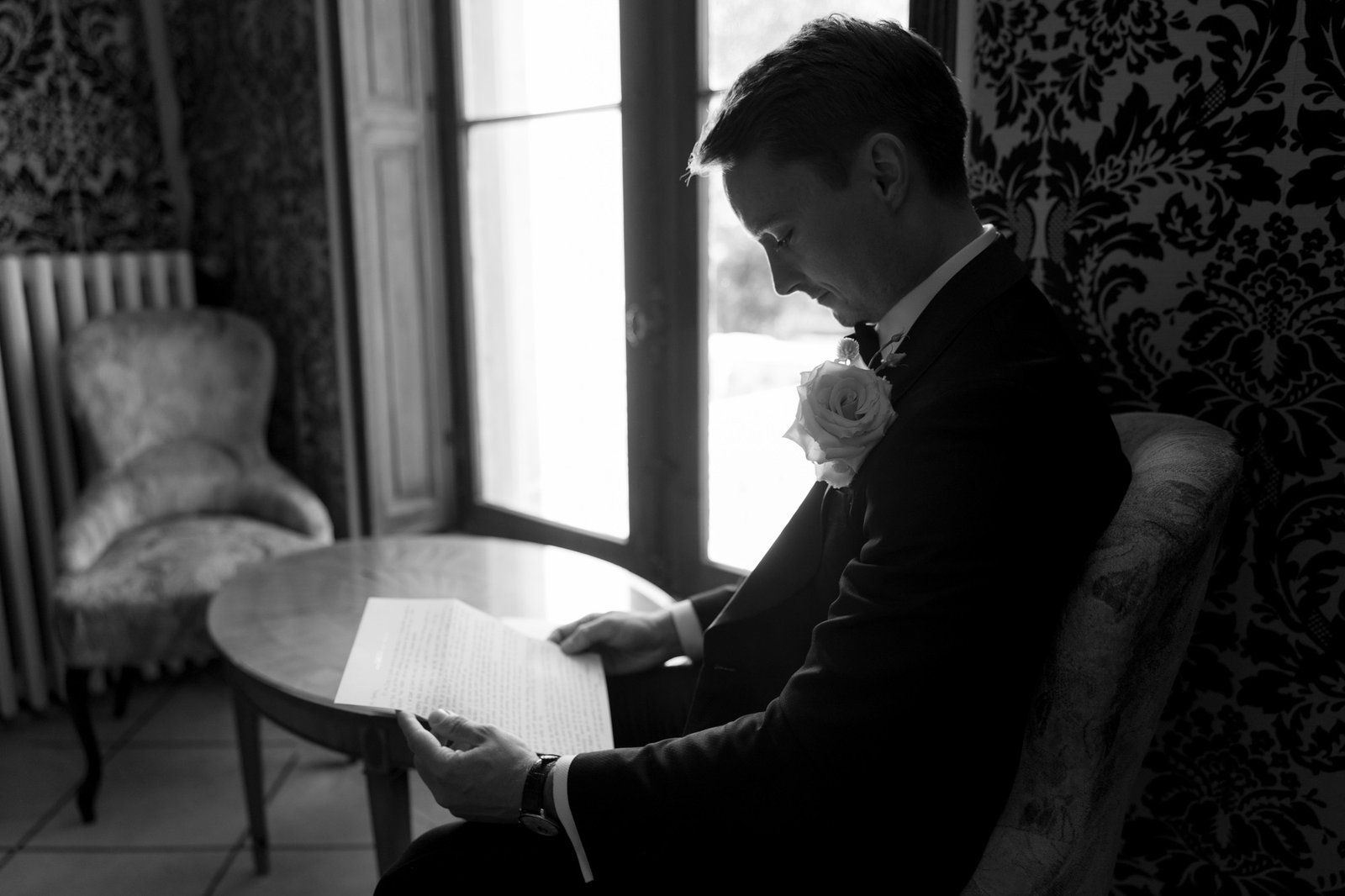 A man in a suit with a boutonnière sits by a window, reading a letter in a dimly lit, ornately decorated room.