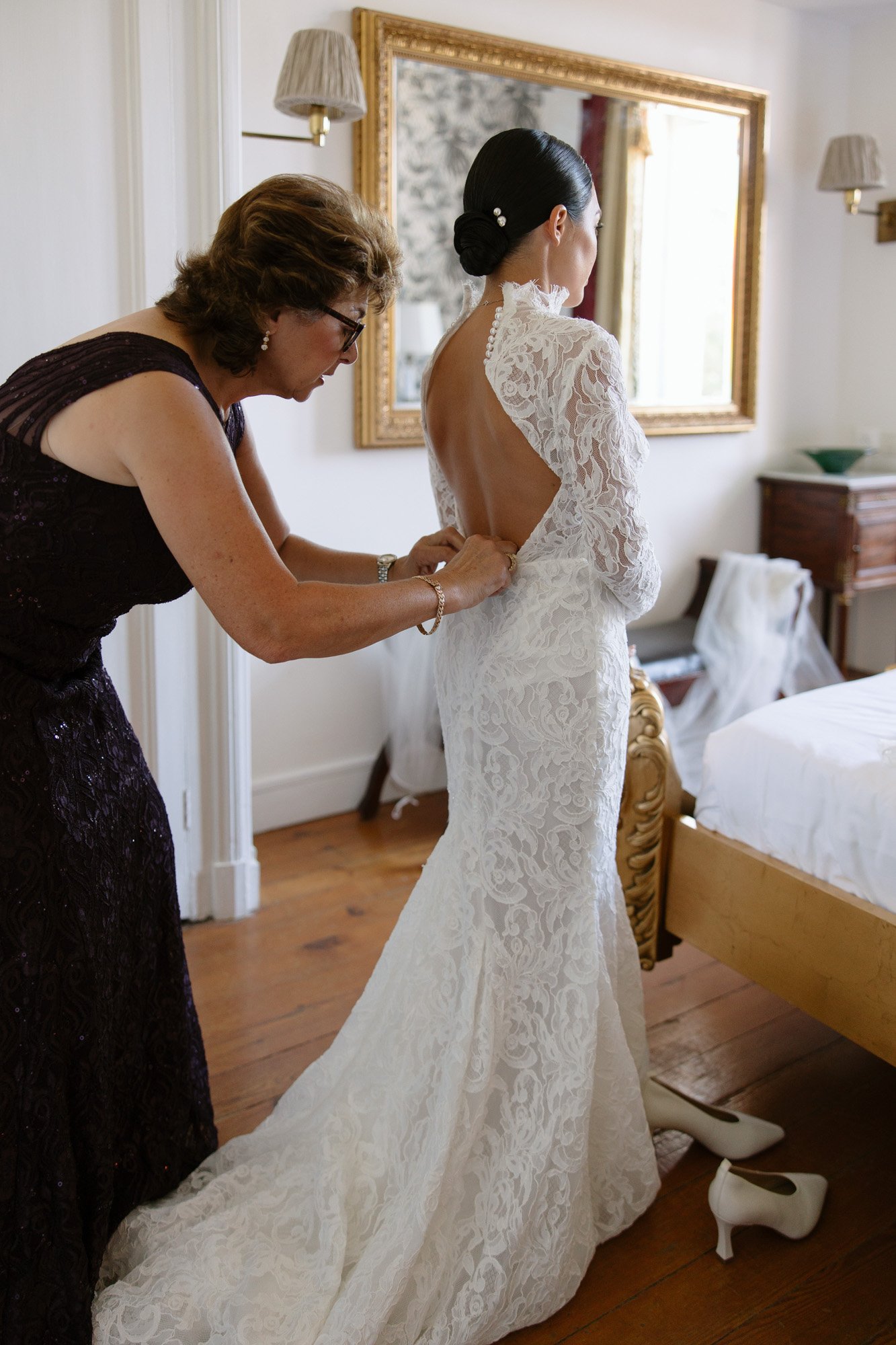 A woman in a lace wedding dress stands as another woman helps fasten the back of her gown in a bedroom with wooden floors and a large mirror.