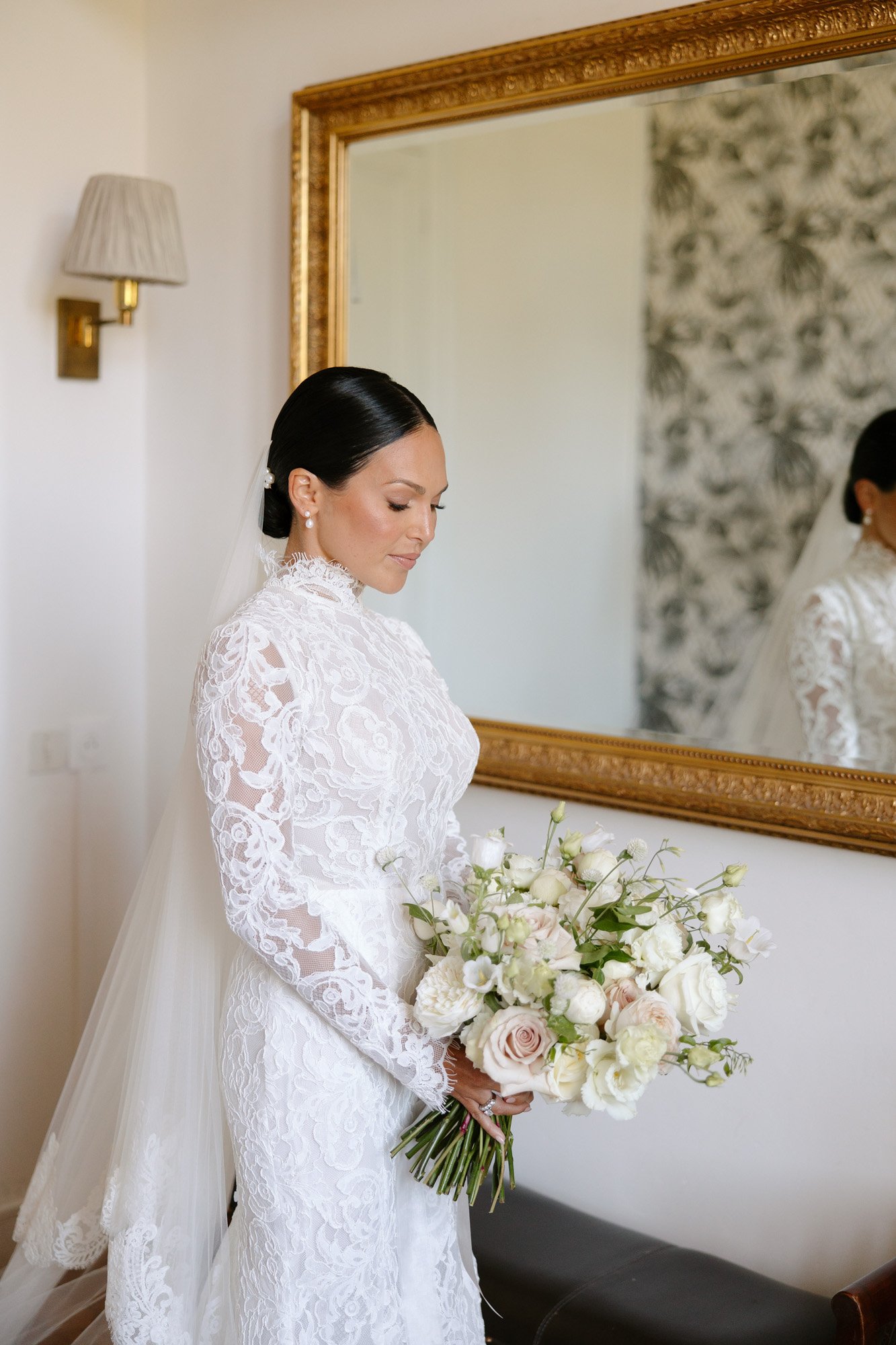A bride in a long-sleeved lace wedding dress holds a bouquet of white and pale pink flowers, standing indoors near a large gold-framed mirror. Chateau de la Valouze wedding.