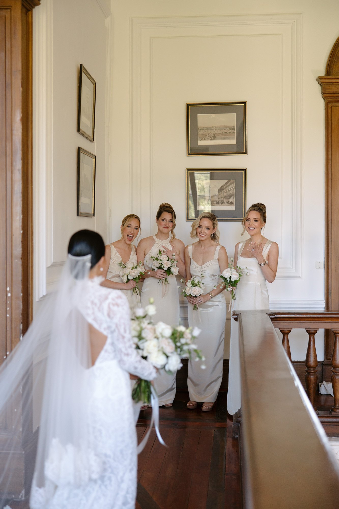 A bride in a white gown and veil approaches four bridesmaids in light dresses, all holding bouquets, standing in a hallway with framed pictures on the wall. Chateau de la Valouze wedding.