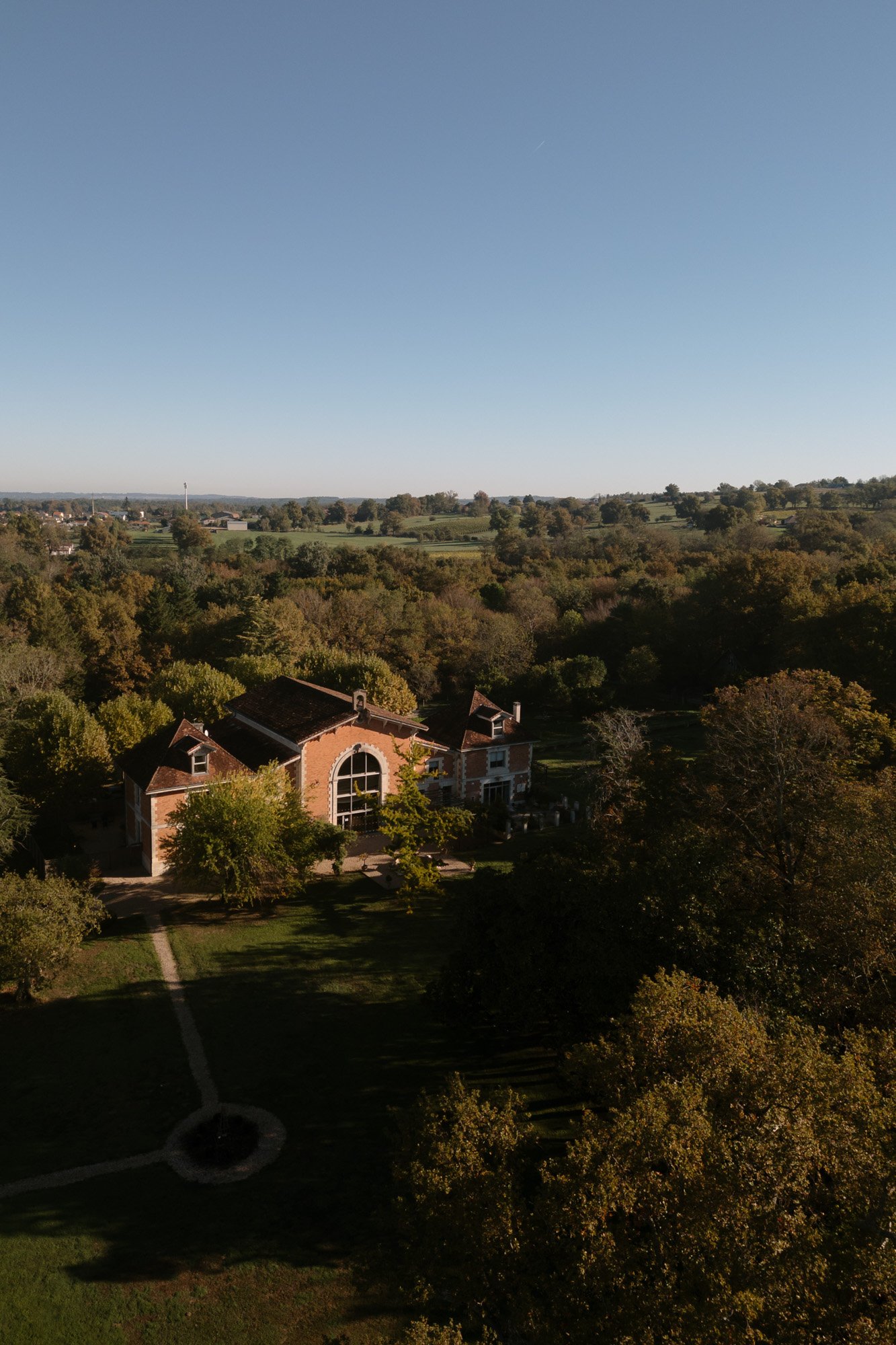 An aerial view of a large brick house with an arched window, surrounded by trees and fields under a clear sky. Chateau de la Valouze wedding.