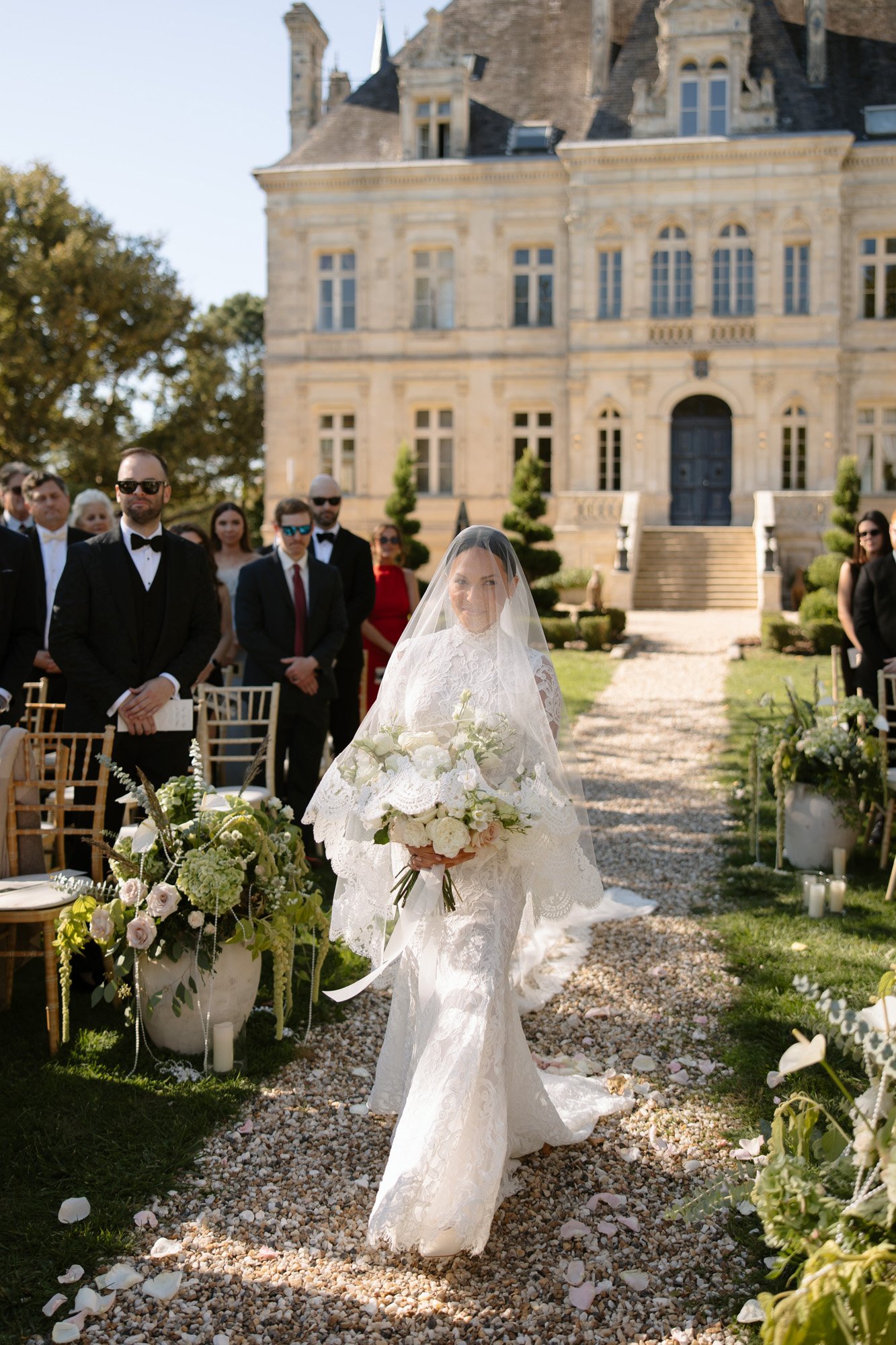 A bride in a white gown and veil walks down an outdoor aisle holding a bouquet, surrounded by seated guests, with a large stone building in the background. Chateau de la Valouze wedding.