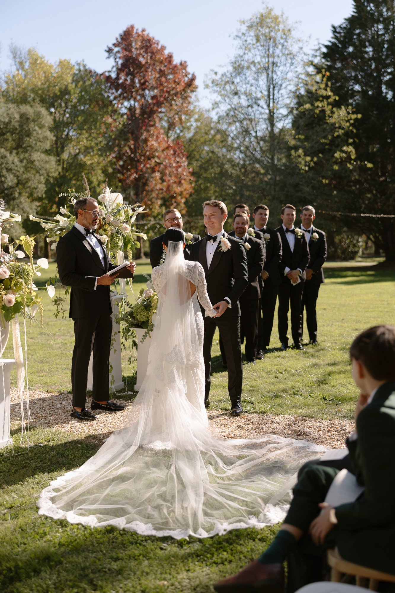 A bride and groom stand at an outdoor wedding altar with a line of groomsmen behind them and a seated guest in the foreground. Chateau de la Valouze wedding.