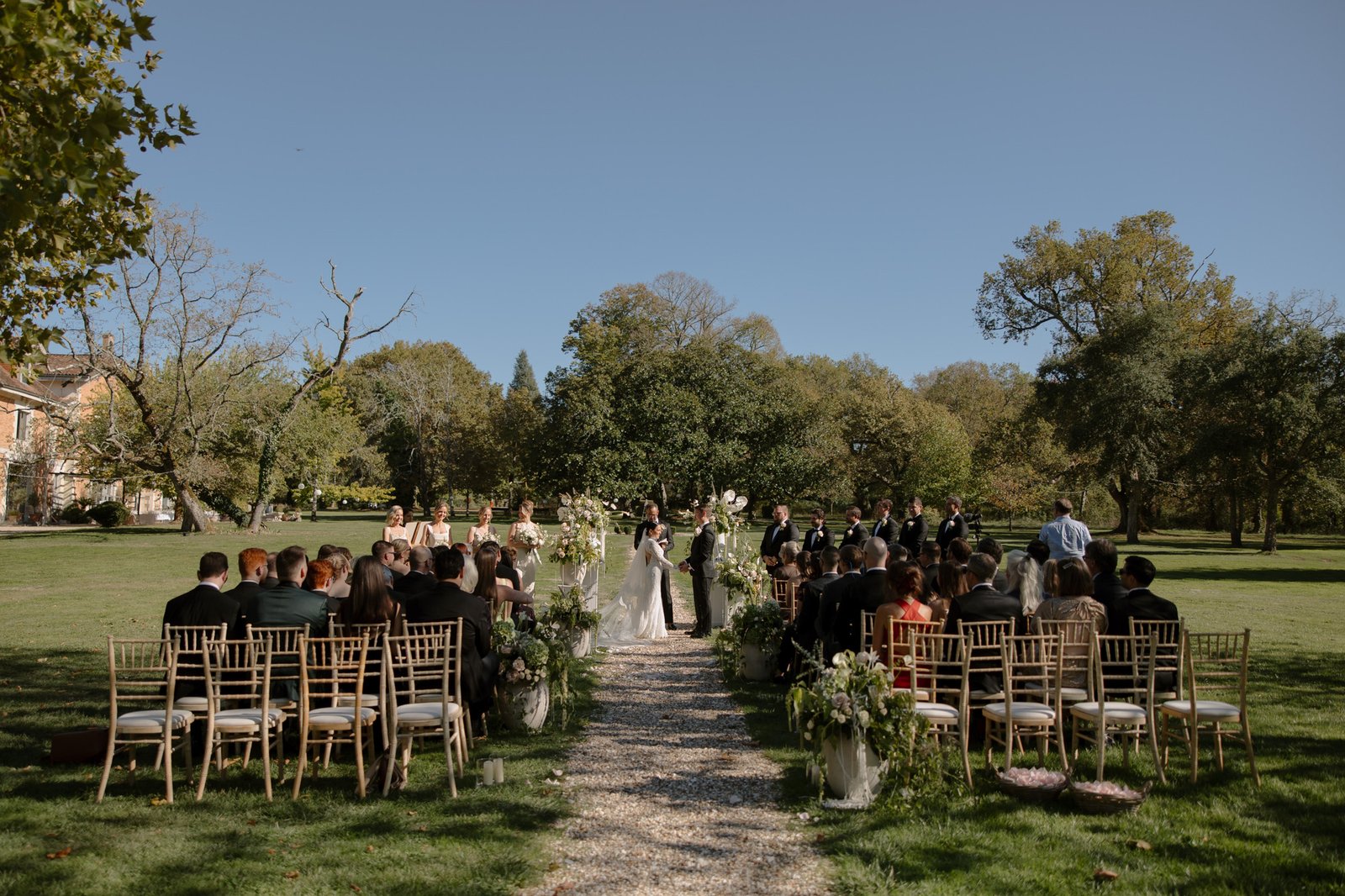 Outdoor wedding ceremony with guests seated on either side of an aisle, bride and groom standing at the altar, surrounded by greenery and trees on a sunny day. Chateau de la Valouze wedding.