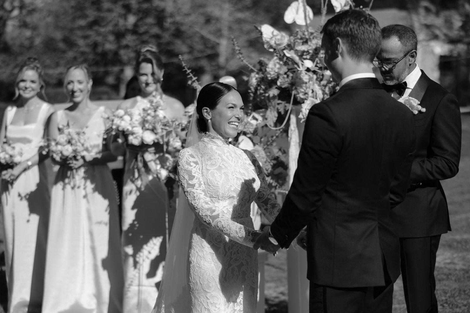 A bride and groom stand facing each other holding hands during an outdoor wedding ceremony, with bridesmaids and an officiant nearby. Chateau de la Valouze wedding.