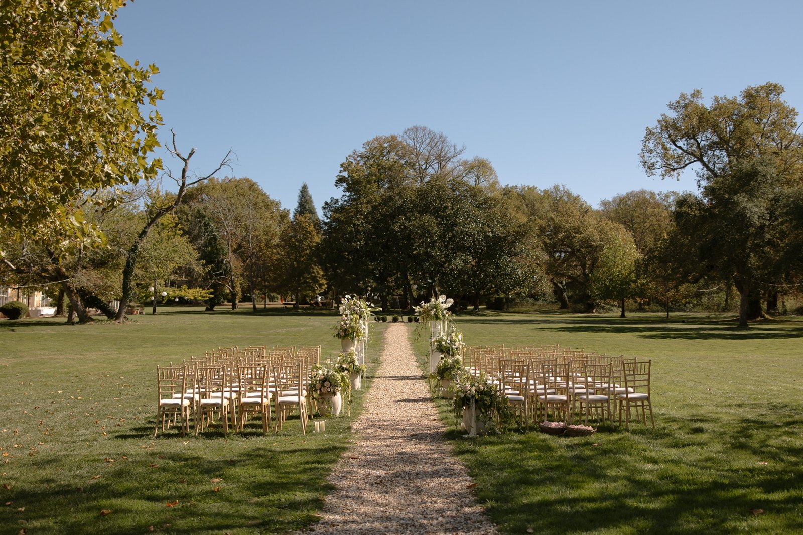 Outdoor wedding ceremony setup with rows of chairs facing an aisle decorated with white flowers, set on a grassy lawn with trees in the background under a clear sky. Chateau de la Valouze wedding.