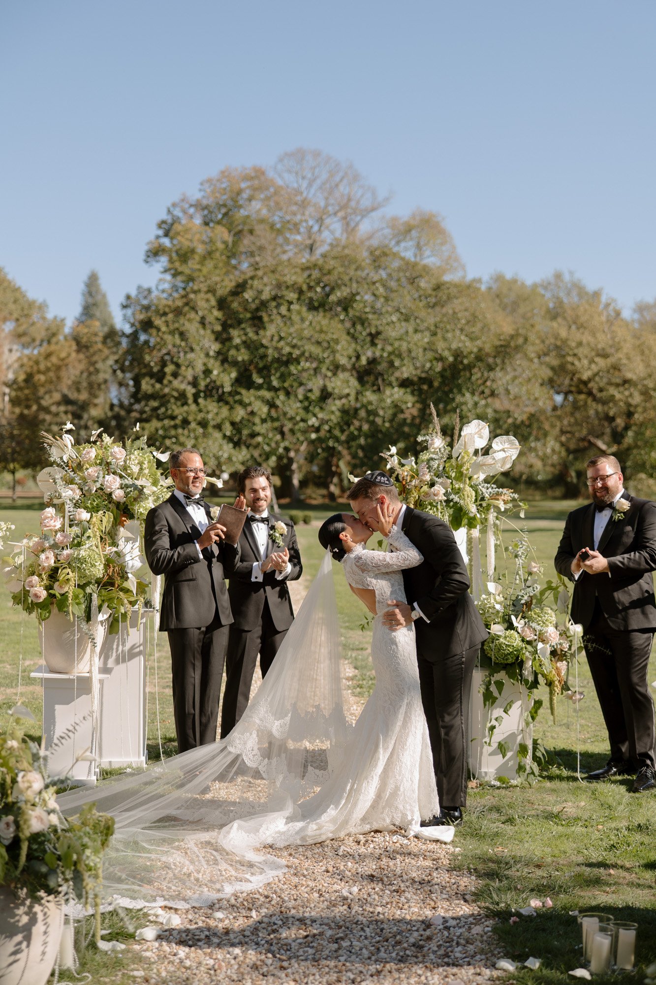 A bride and groom kiss at an outdoor wedding ceremony while groomsmen stand nearby, surrounded by floral arrangements on a sunny day.