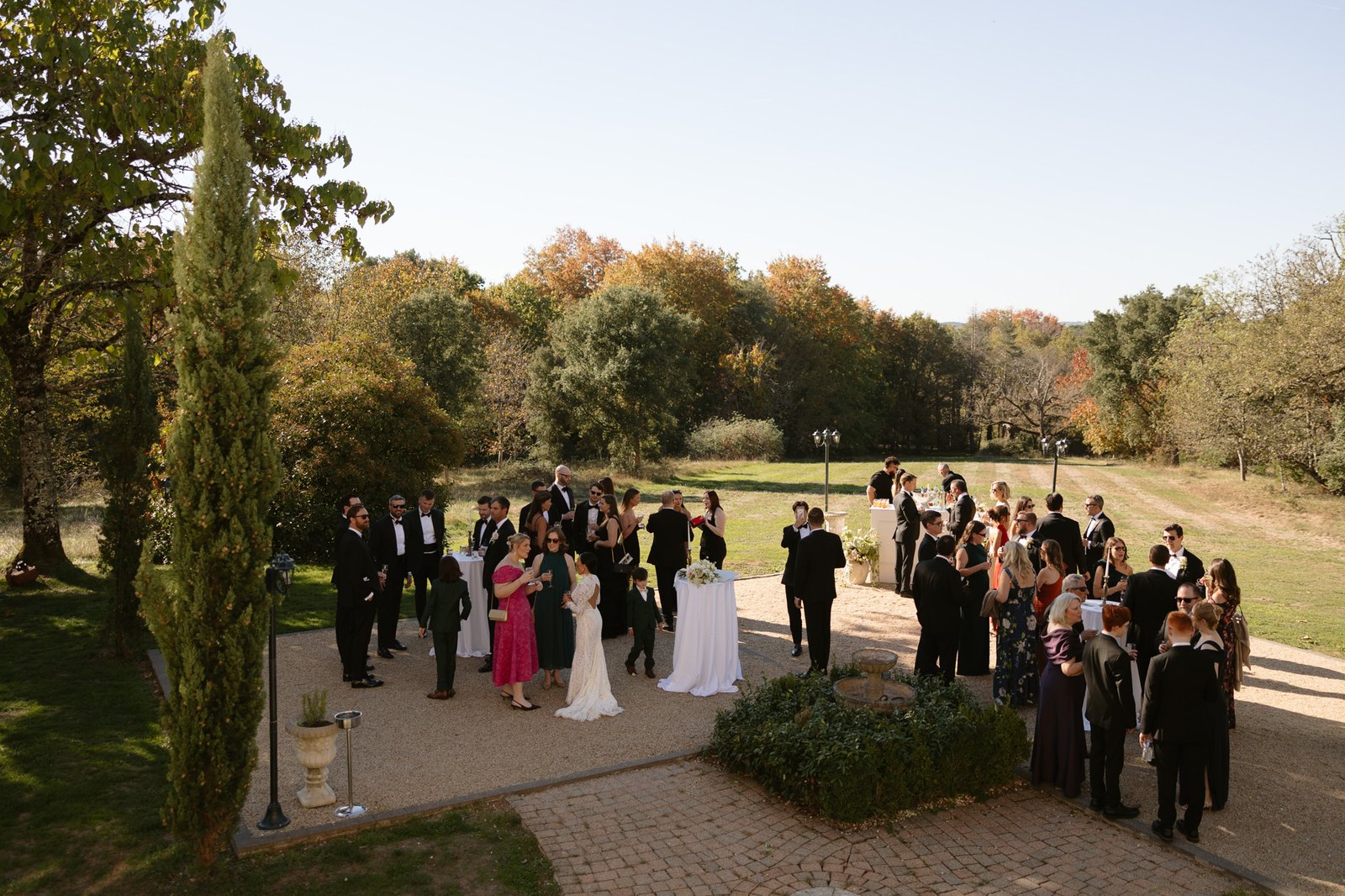 A group of people dressed formally gather outdoors on a sunny day, standing in small groups near cocktail tables, surrounded by trees and greenery. Chateau de la Valouze wedding.