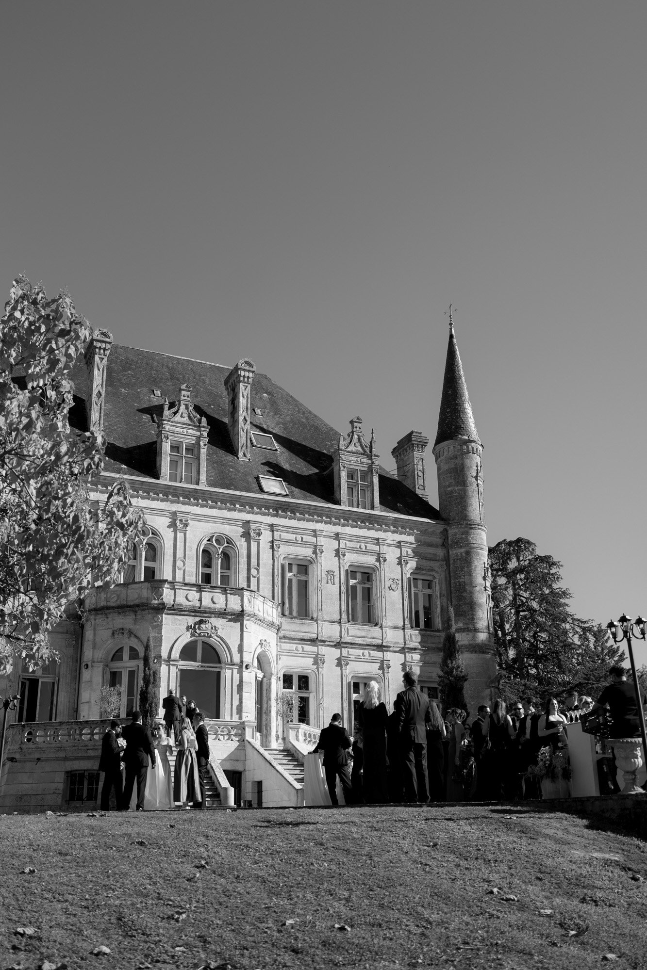 A group of people gathers outside a large, ornate stone building with a turret under a clear sky. Chateau de la Valouze wedding.