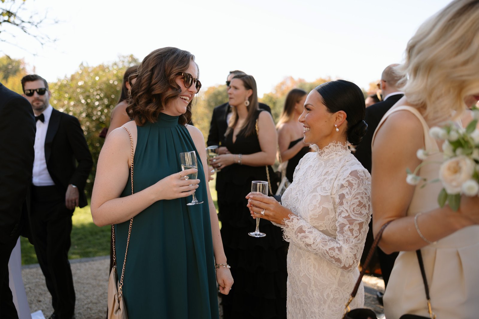 Two women, one in a white lace dress and the other in a green dress, stand outdoors holding champagne glasses and talking at a formal event.