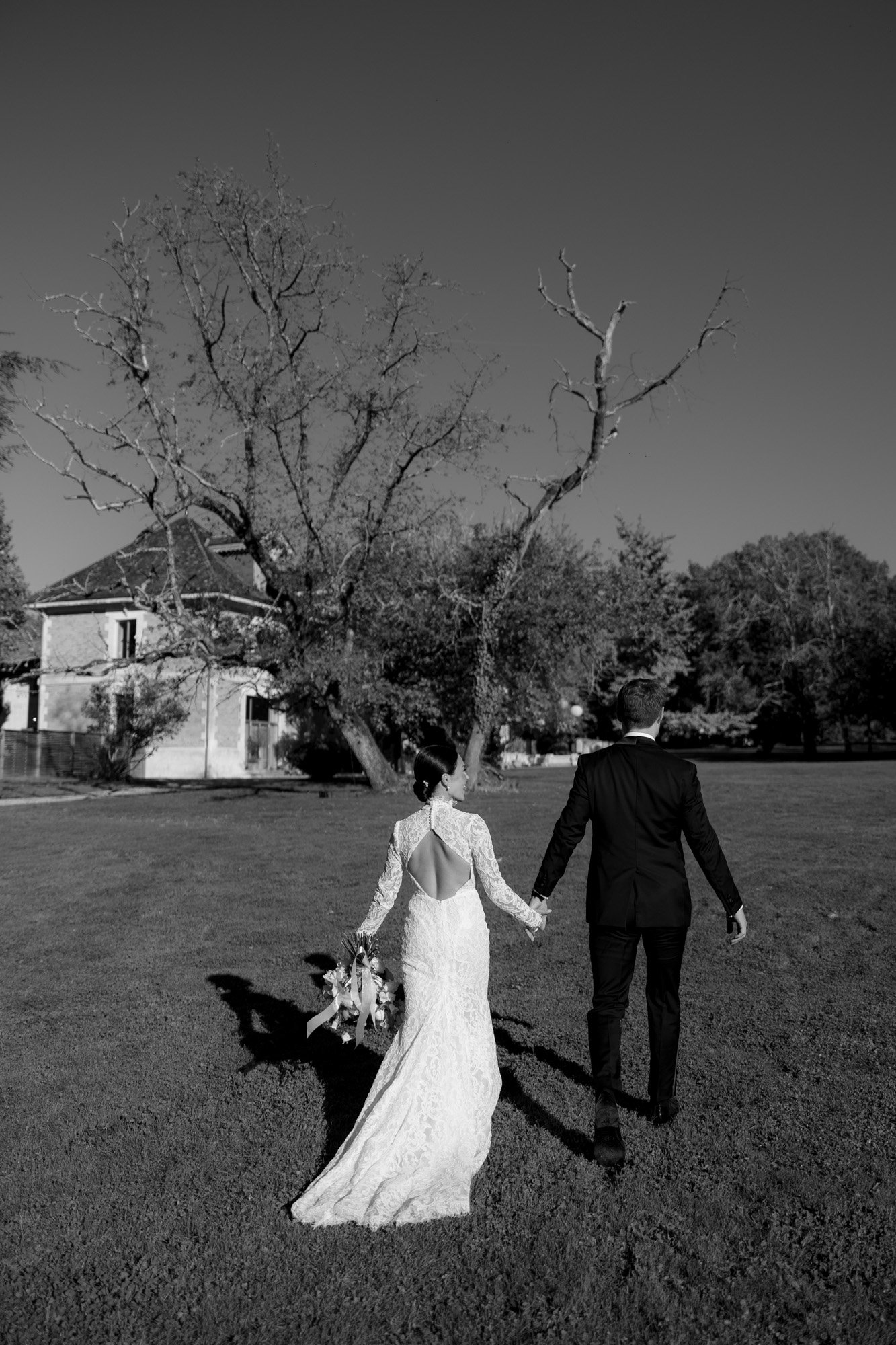 A bride and groom walk hand in hand across a lawn toward a leafless tree, with a house visible in the background. Chateau de la Valouze wedding.