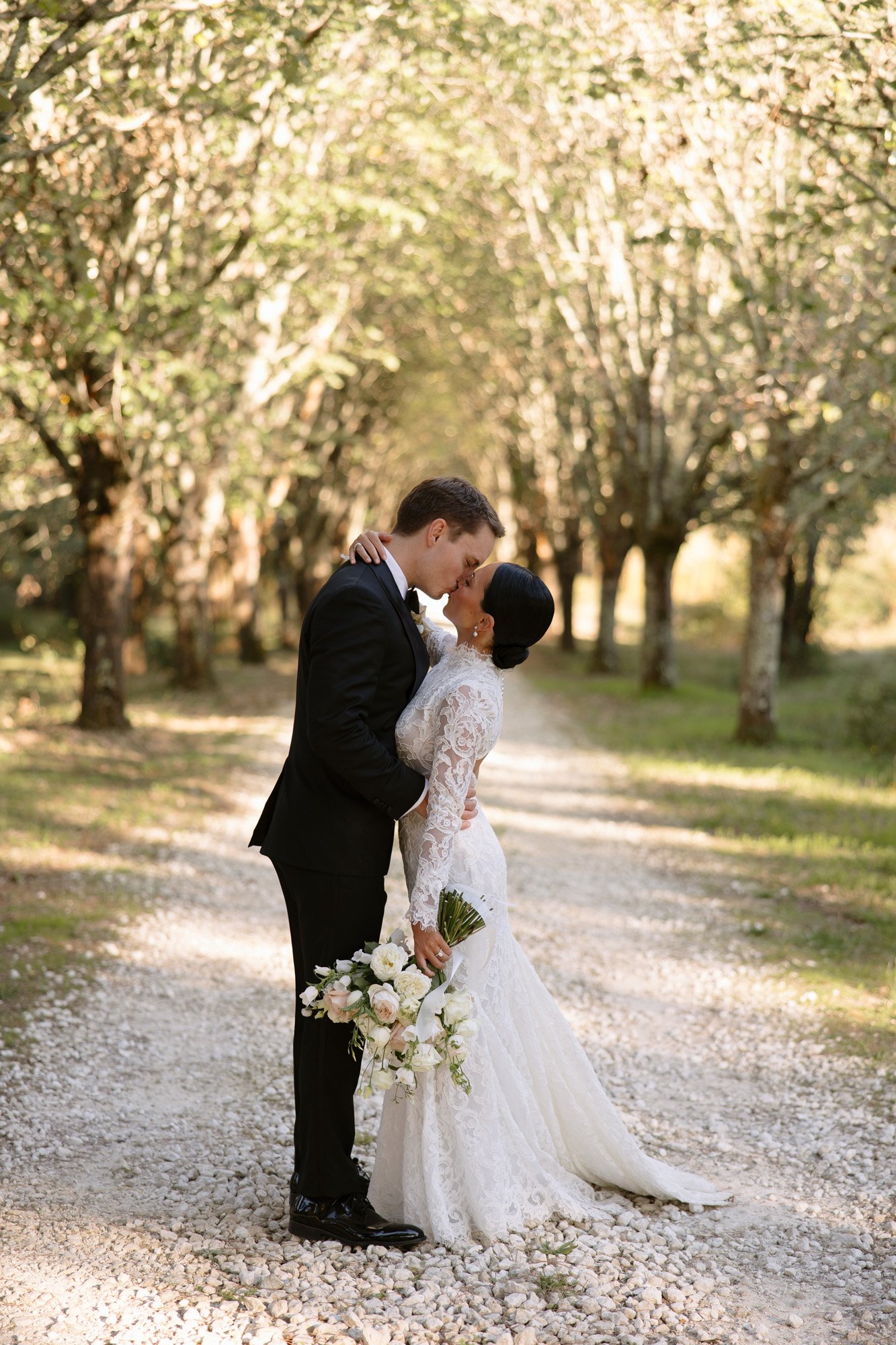 A bride and groom in formal wedding attire share a kiss on a tree-lined gravel path, with the bride holding a bouquet of white flowers. Chateau de la Valouze wedding.