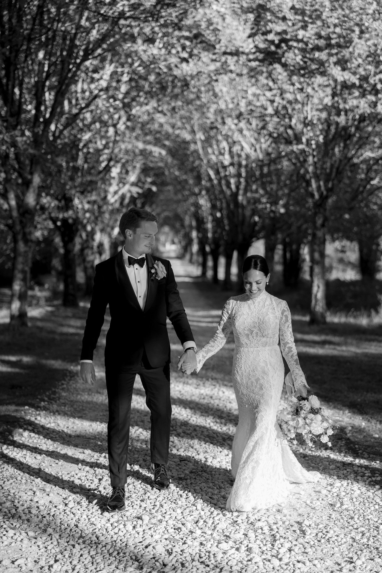 A bride and groom walk hand in hand down a tree-lined path, with the bride holding a bouquet and both dressed in formal wedding attire. Chateau de la Valouze wedding.