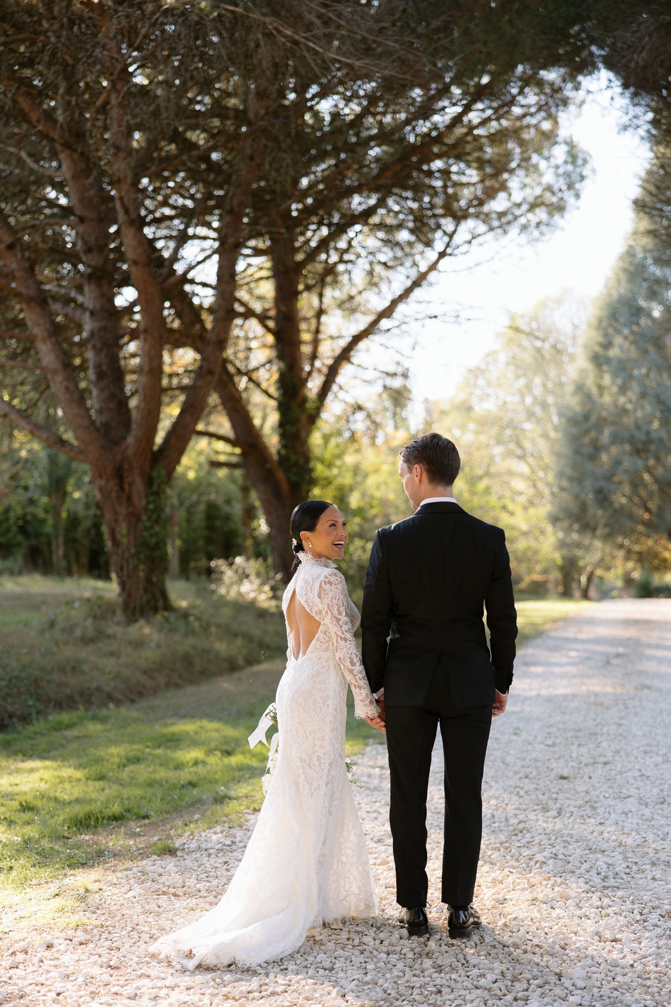A bride in a white lace gown and a groom in a black suit stand on a gravel path outdoors, holding hands and looking at each other, surrounded by trees. Chateau de la Valouze wedding.