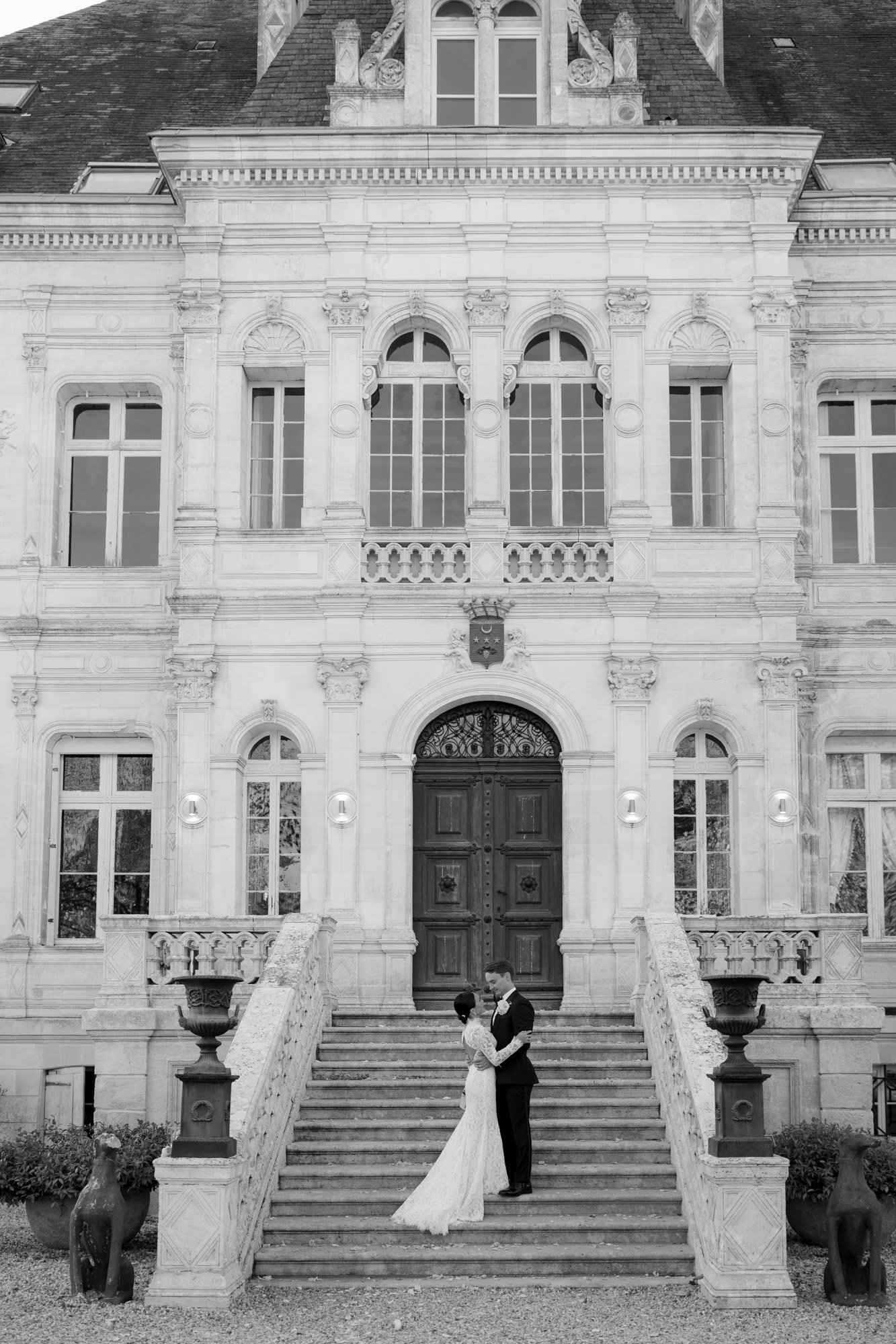 A bride and groom stand embracing on the steps of a grand, ornate building with large doors and arched windows. Chateau de la Valouze wedding.