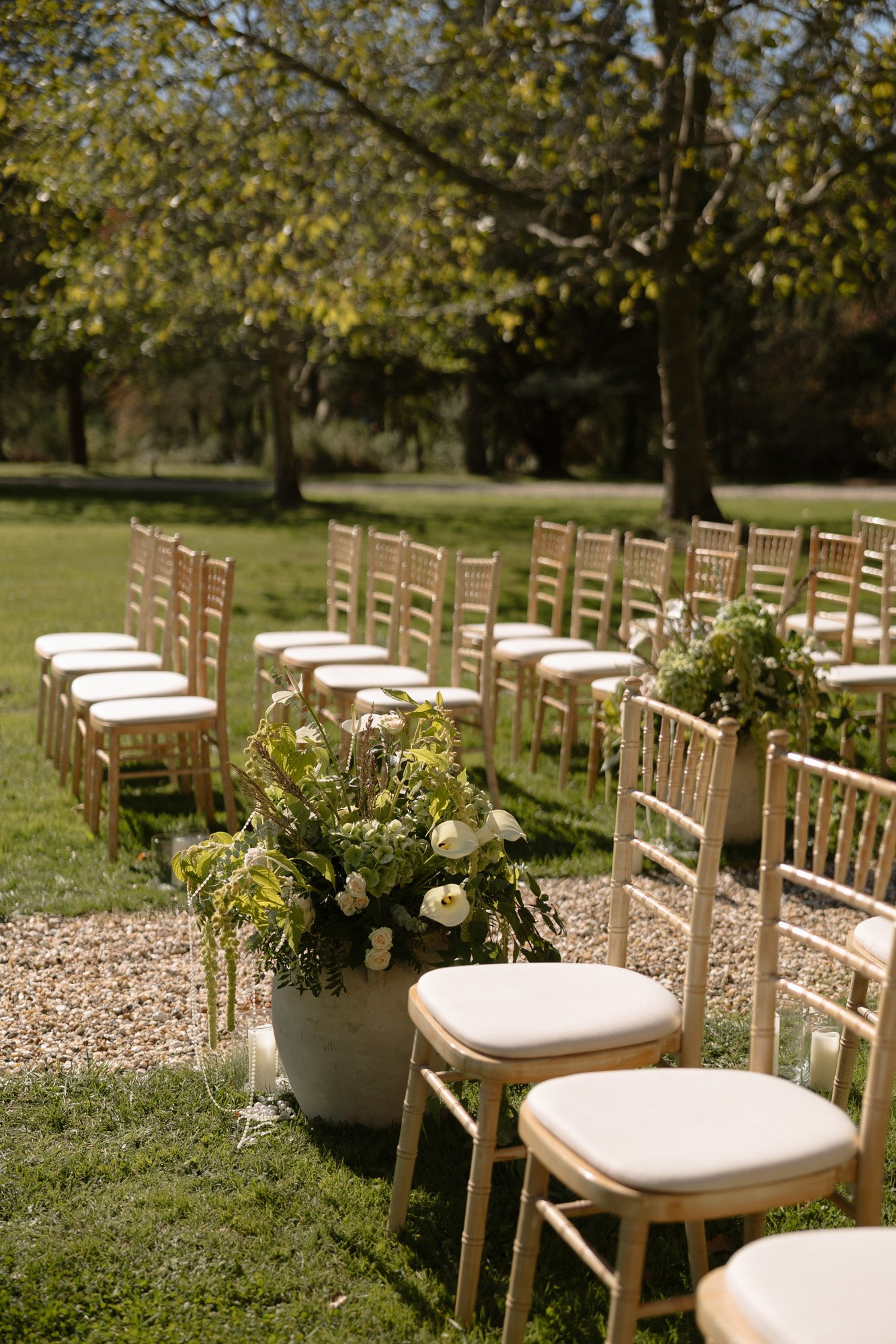 Rows of wooden chairs are arranged outdoors on a grassy lawn, with planters of white flowers and greenery placed at the aisle.