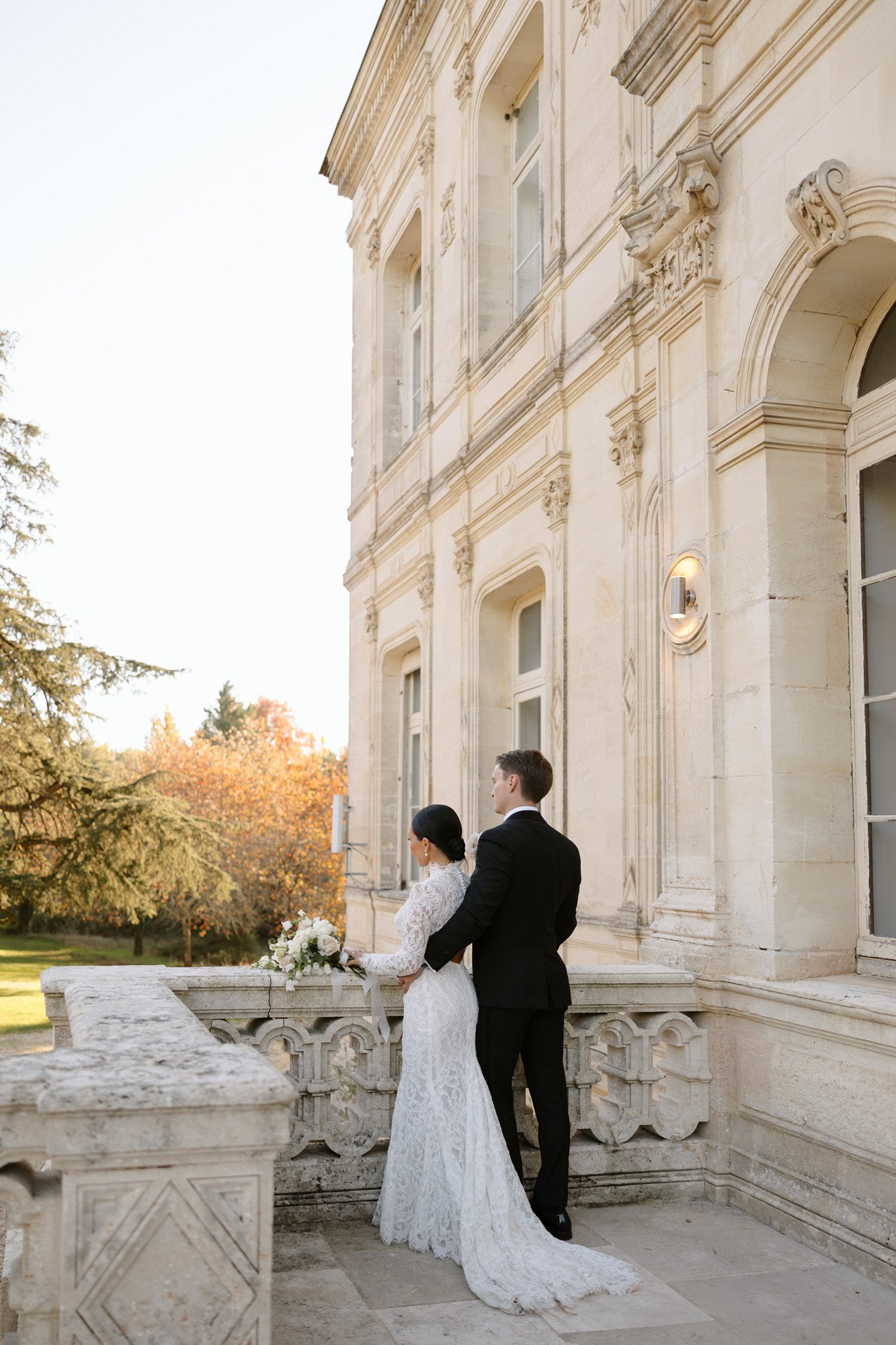 A bride and groom stand on a stone balcony, facing away, overlooking a garden outside a light-colored historic building. Chateau de la Valouze wedding.
