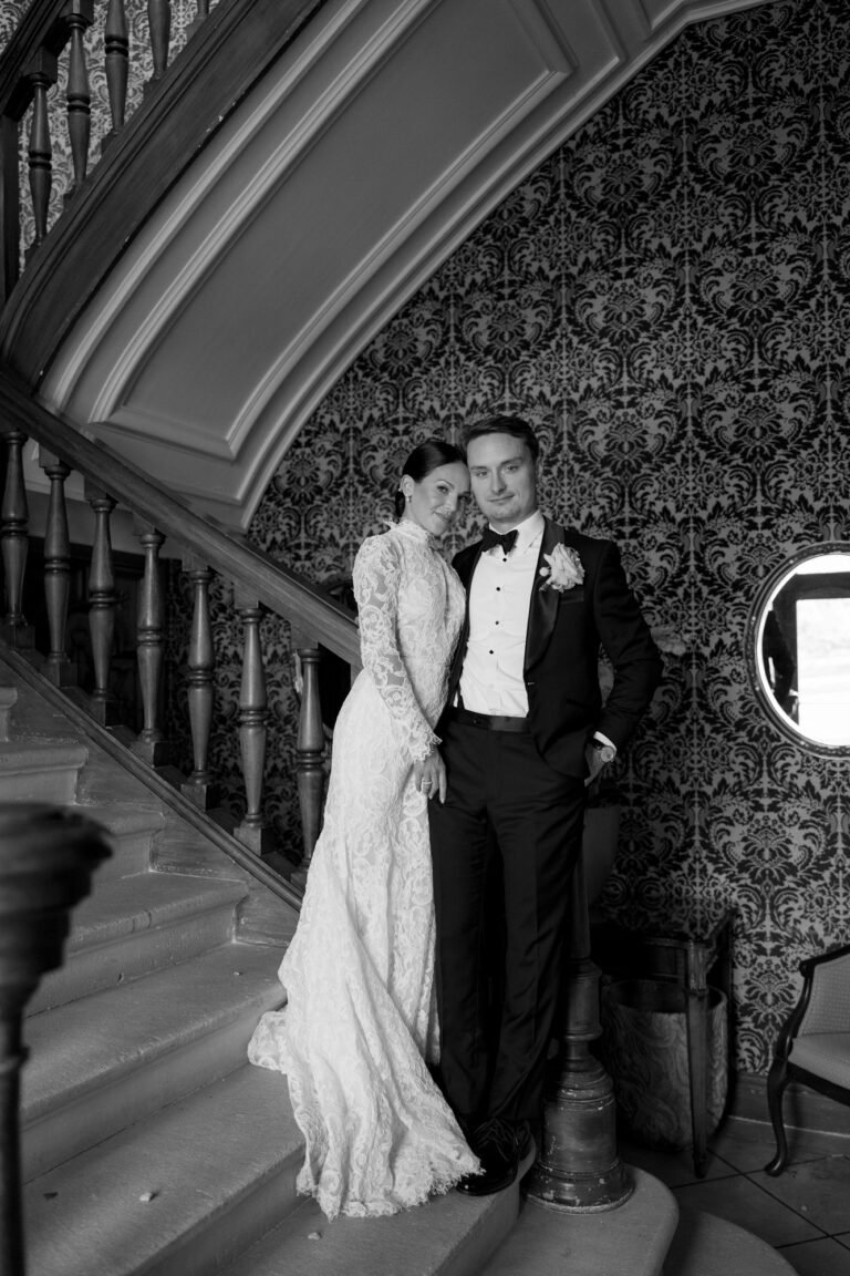 A bride in a long lace gown and a groom in a tuxedo pose together on a staircase beneath ornate wallpaper, both looking at the camera.