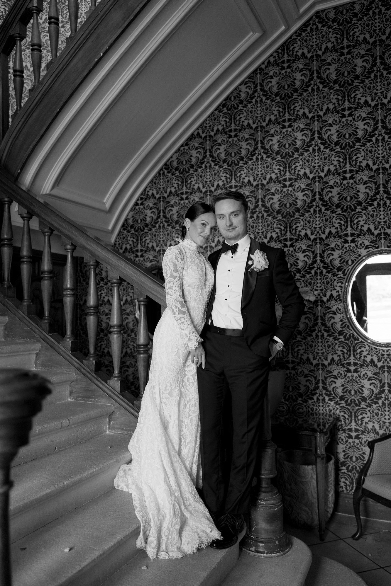 A bride in a long lace gown and a groom in a tuxedo pose together on a staircase beneath ornate wallpaper, both looking at the camera.