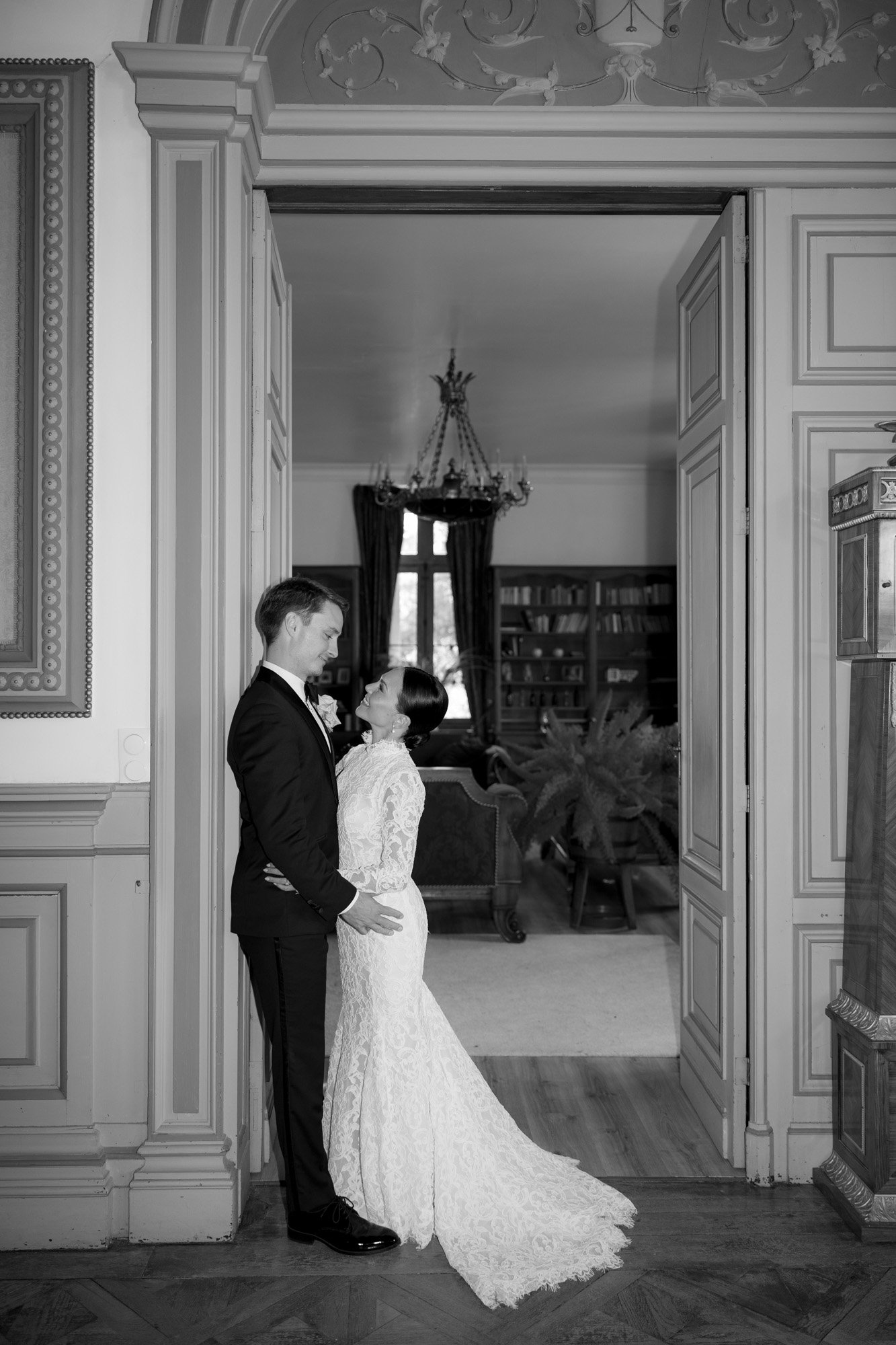 A couple in wedding attire stands facing each other in a doorway of an ornate room, the bride in a lace dress and the groom in a suit, with a chandelier and bookshelves in the background. Chateau de la Valouze wedding.