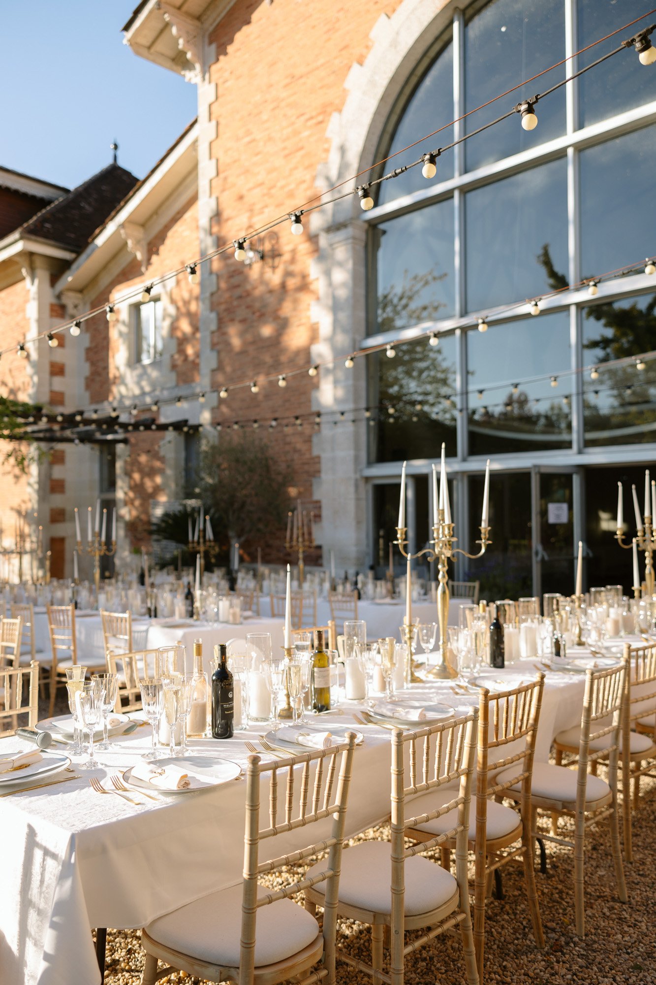 Outdoor event setup with white-clothed tables, elegant place settings, candelabras, and string lights in front of a large brick building with tall windows. Chateau de la Valouze wedding.