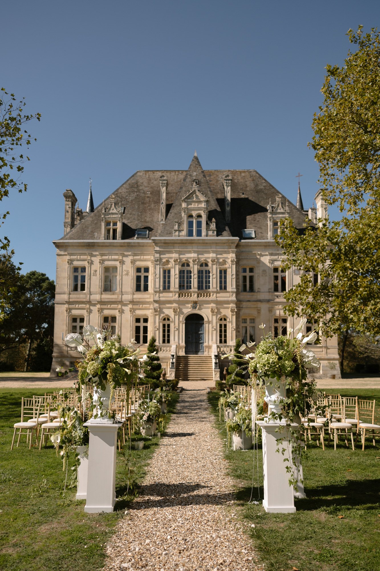 A stone path lined with floral arrangements leads to an outdoor wedding setup in front of a large historic chateau on a clear day. Chateau de la Valouze wedding.