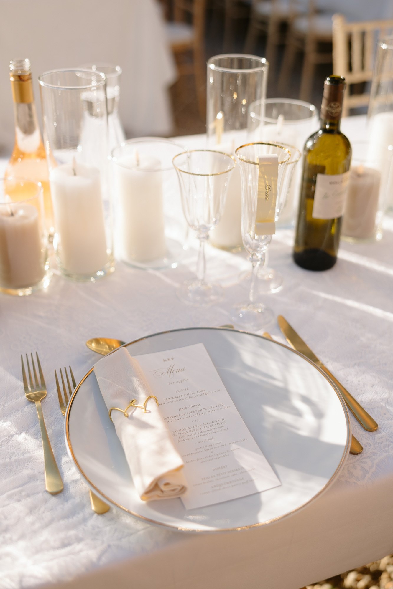 Elegant table setting with a gold-rimmed plate, menu, rolled napkin, cutlery, wine glasses, bottles, and white candles on a white tablecloth. Chateau de la Valouze wedding.