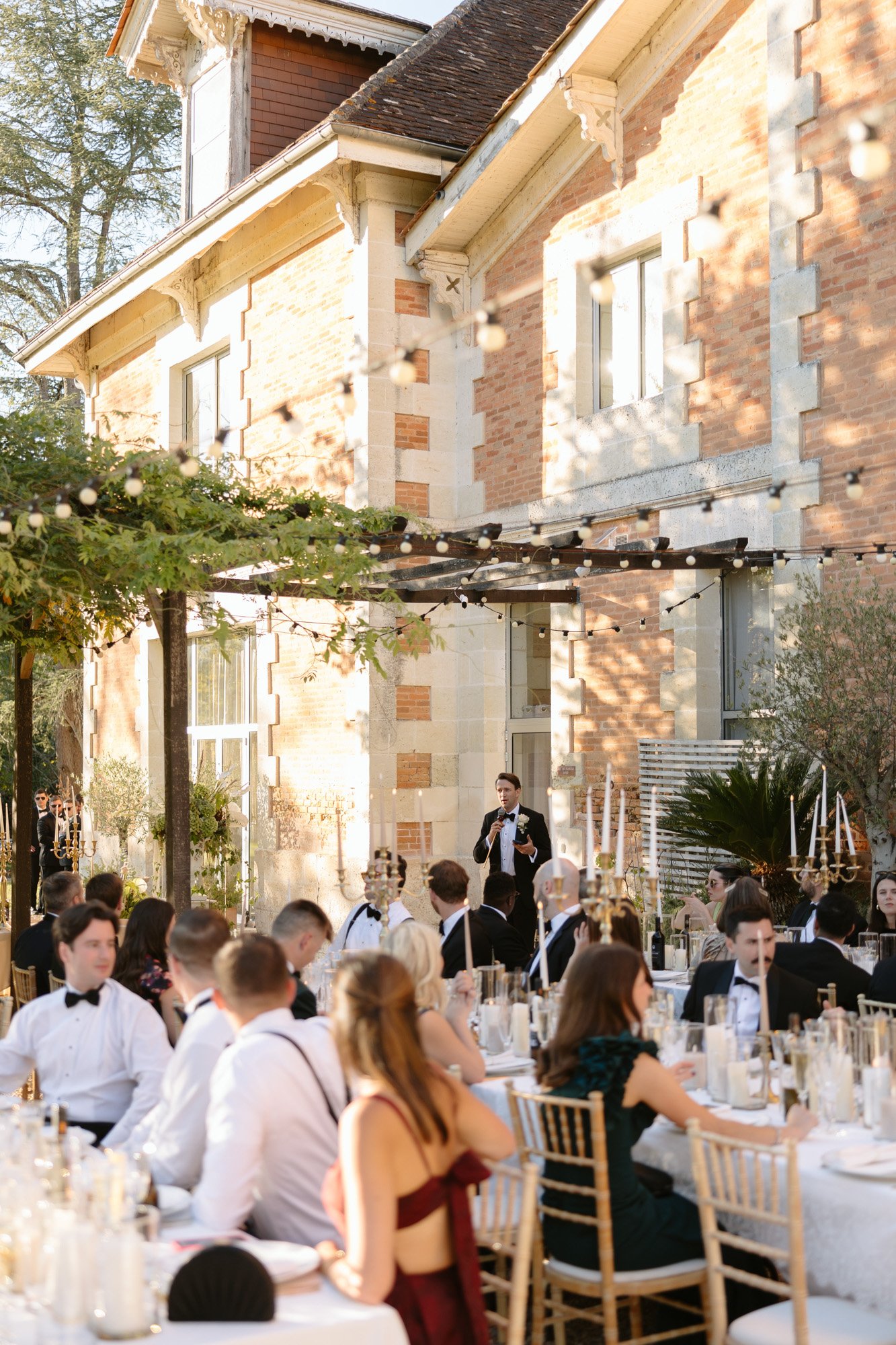 A group of people seated at outdoor tables for a formal event; a man stands and speaks into a microphone in front of a brick building decorated with string lights.