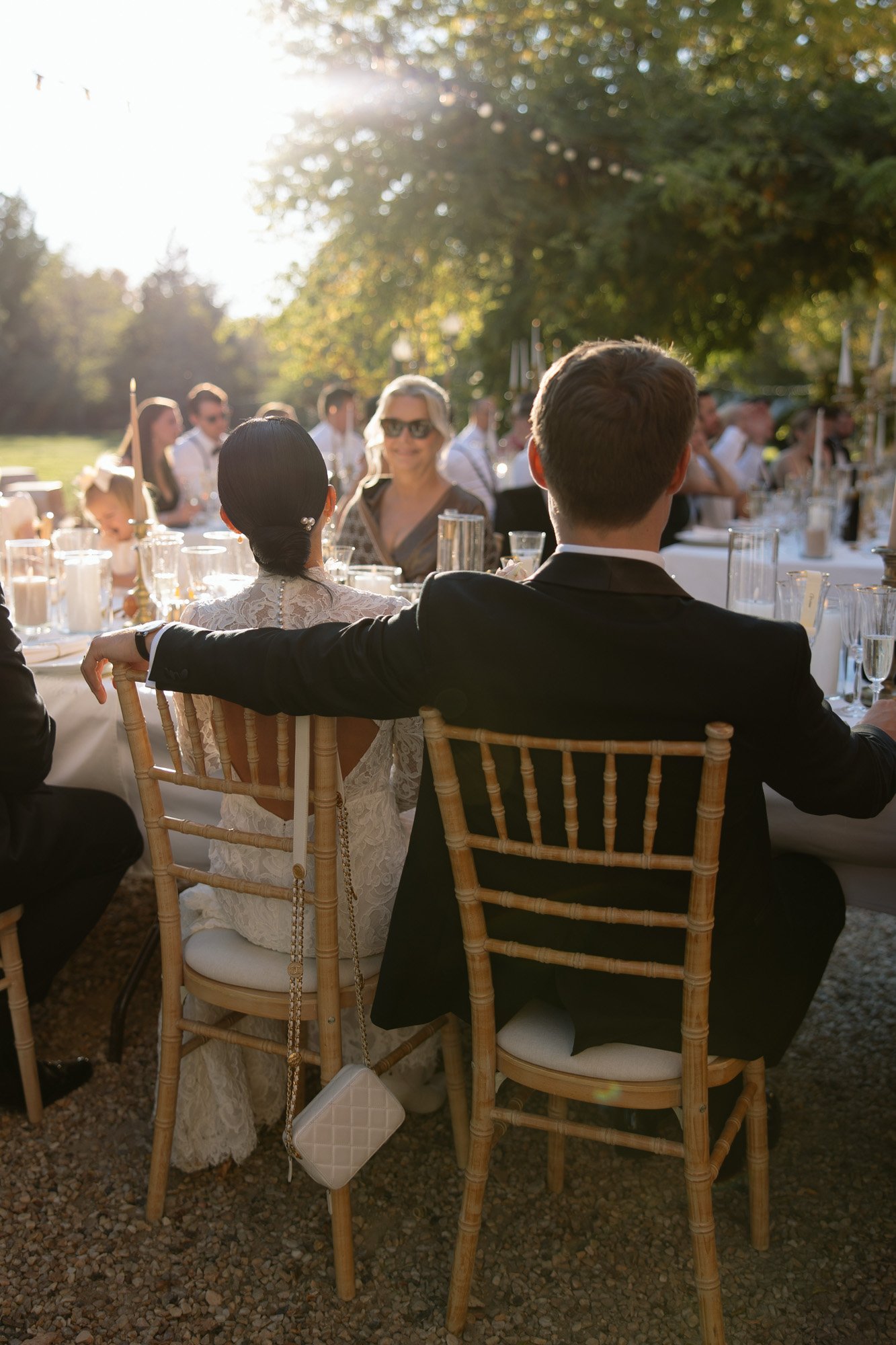 A couple sits closely together at an outdoor event, facing a table with guests and glassware, in soft sunlight. Chateau de la Valouze wedding.