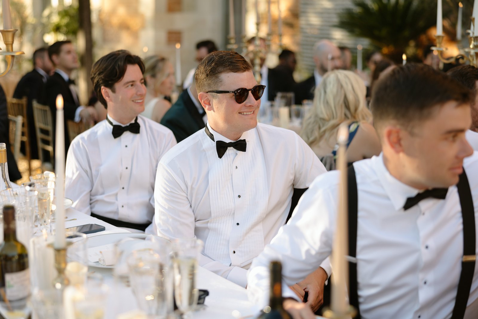 Three men in tuxedos sit at a formal outdoor event table with candles, glasses, and wine bottles, surrounded by other guests.