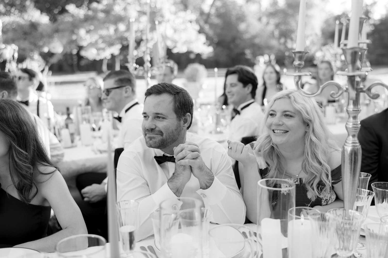 A group of people dressed formally sit at a long outdoor table set with candles and glasses, appearing to listen attentively during a daytime event.
