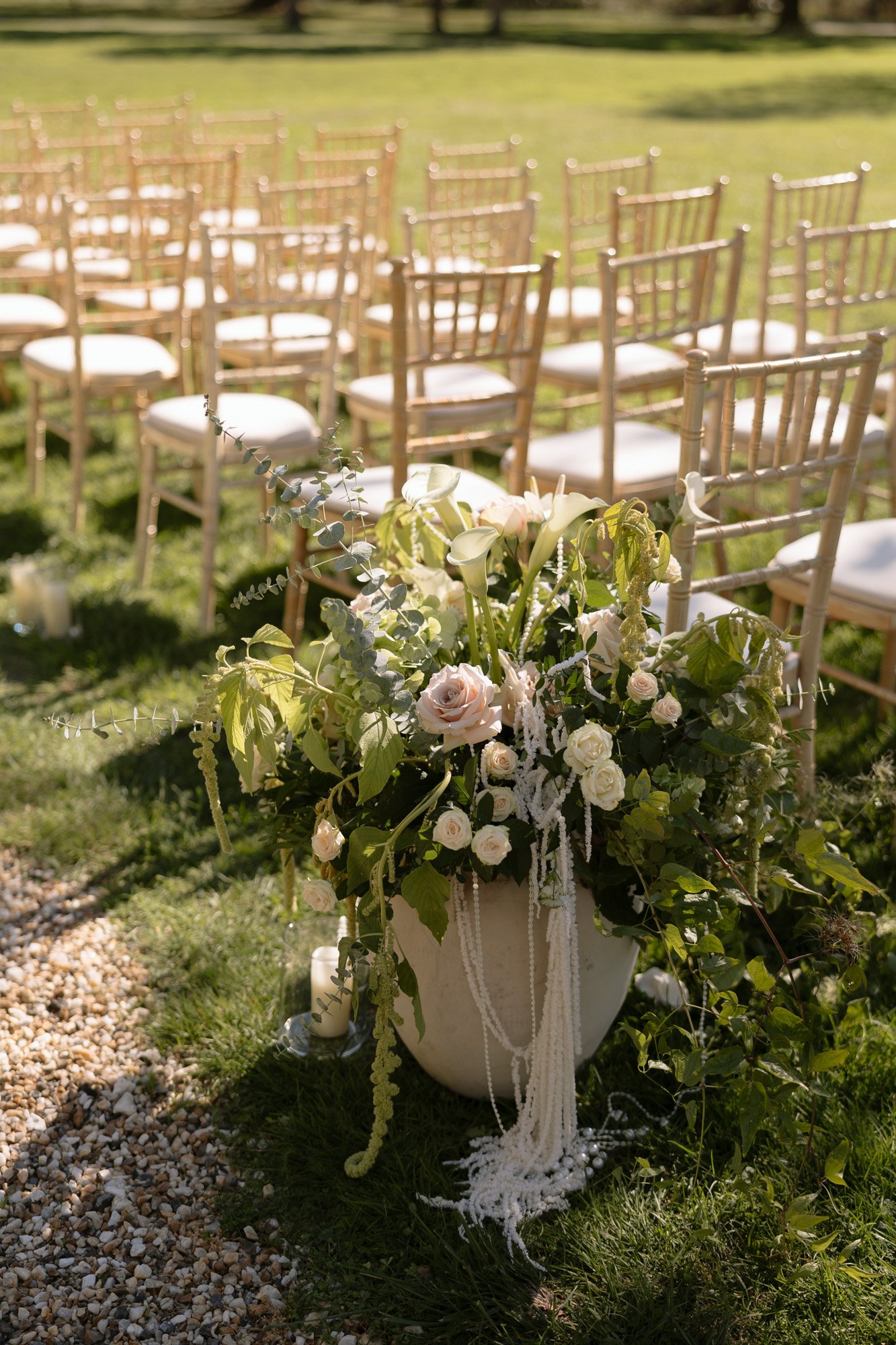 Rows of wooden chairs arranged outdoors on grass, with a large floral arrangement of white and pale pink flowers and greenery at the aisle end. Chateau de la Valouze wedding.