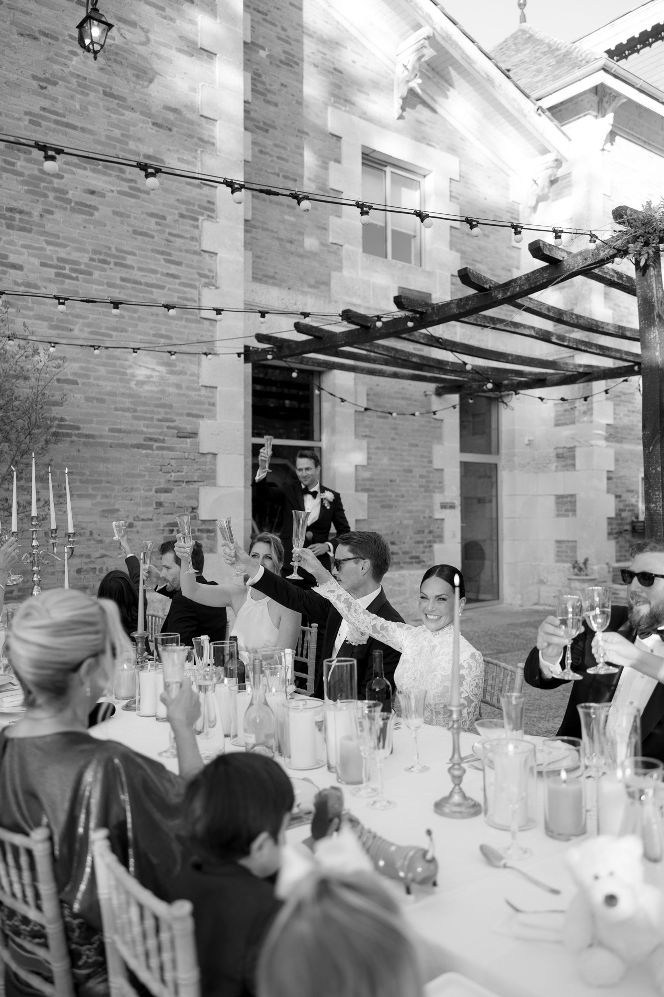A group of people dressed formally sit around a long outdoor table, raising glasses in a toast, in front of a stone building. Chateau de la Valouze wedding.