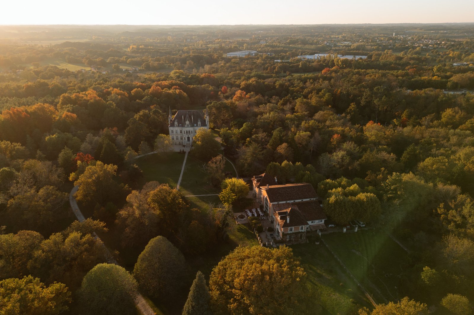 Aerial view of two large buildings surrounded by dense forest and autumn foliage, with a distant horizon under soft sunlight. Chateau de la Valouze wedding.