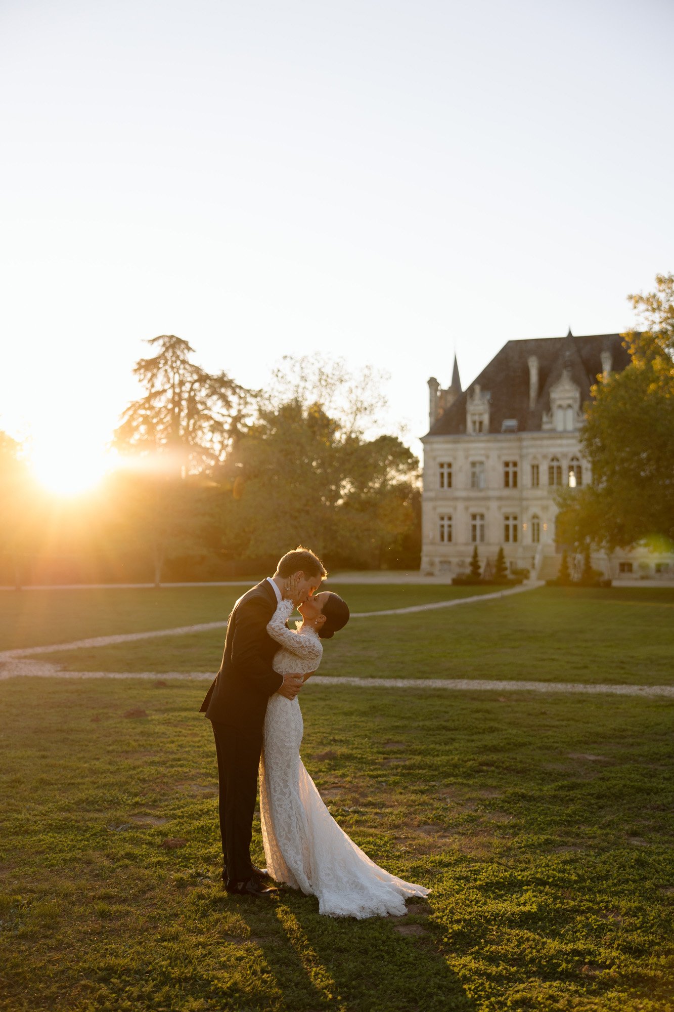 A bride and groom embrace and kiss outdoors at sunset, with a large historic building and trees in the background. Chateau de la Valouze wedding.
