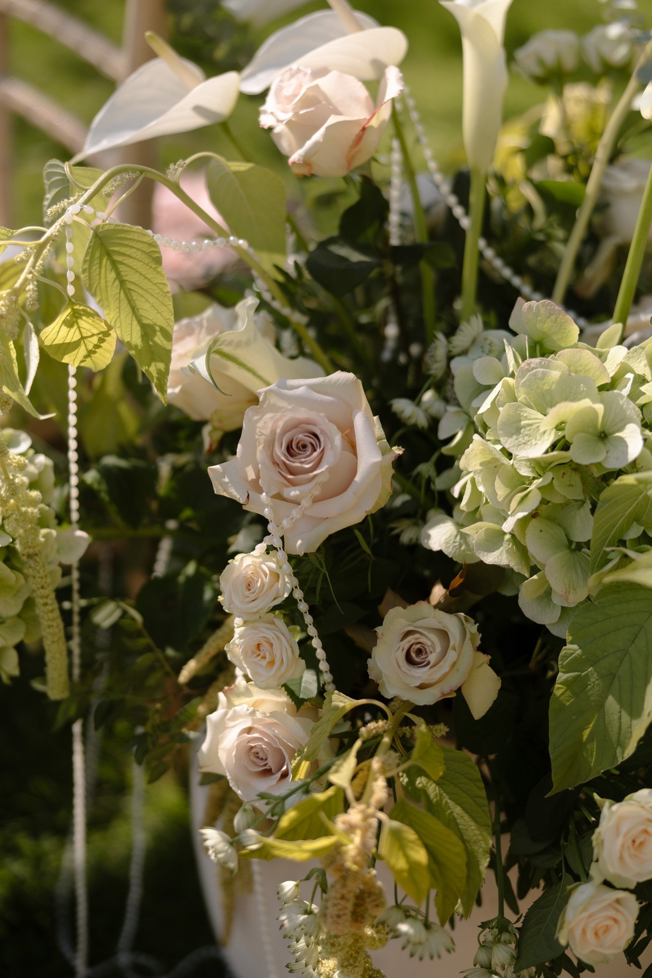 A floral arrangement featuring pale roses, white calla lilies, green hydrangeas, leaves, and strands of white beads.
