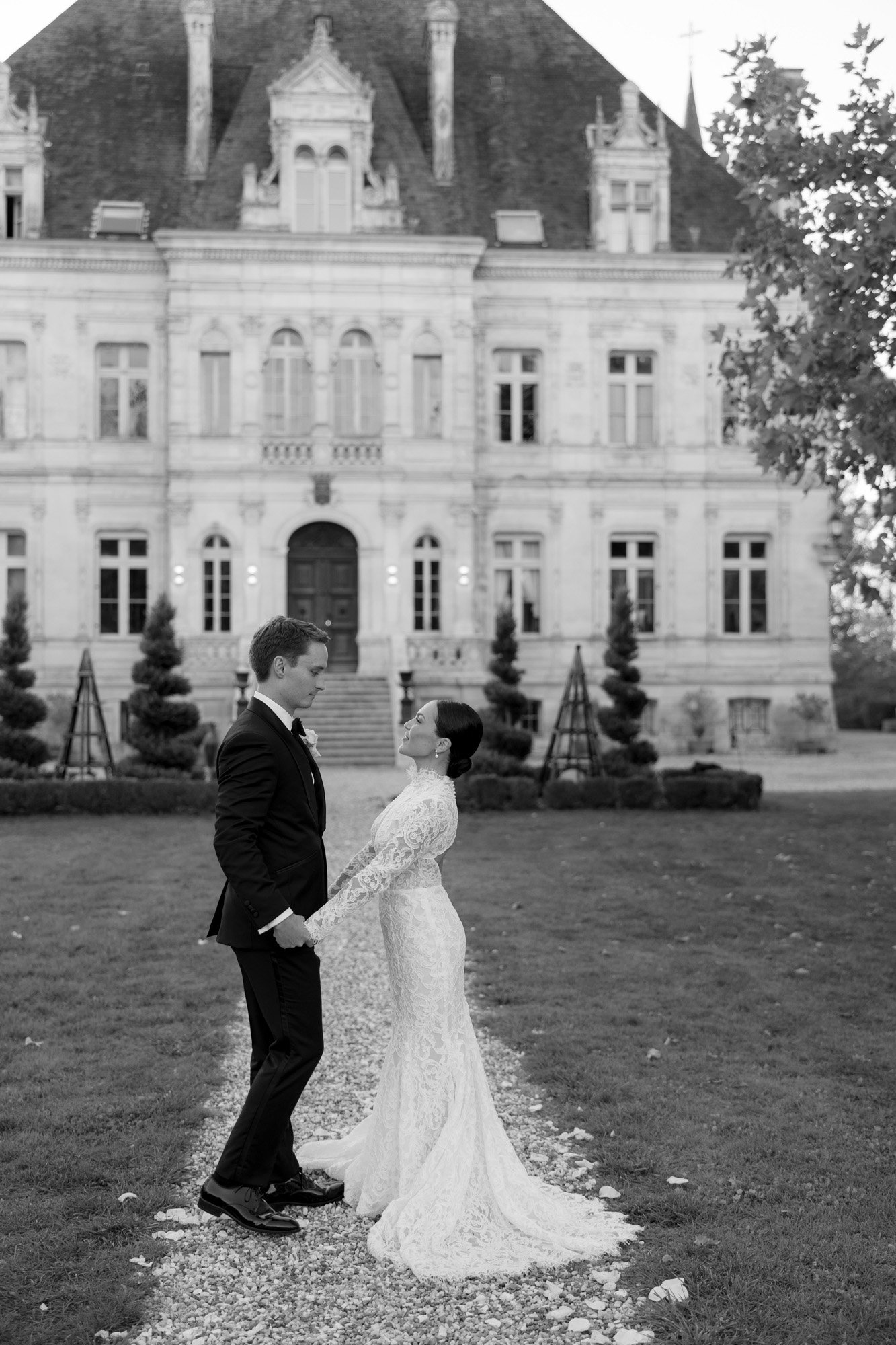 A bride and groom stand facing each other, holding hands, on a path outside a large historic building with ornate architecture. Chateau de la Valouze wedding.
